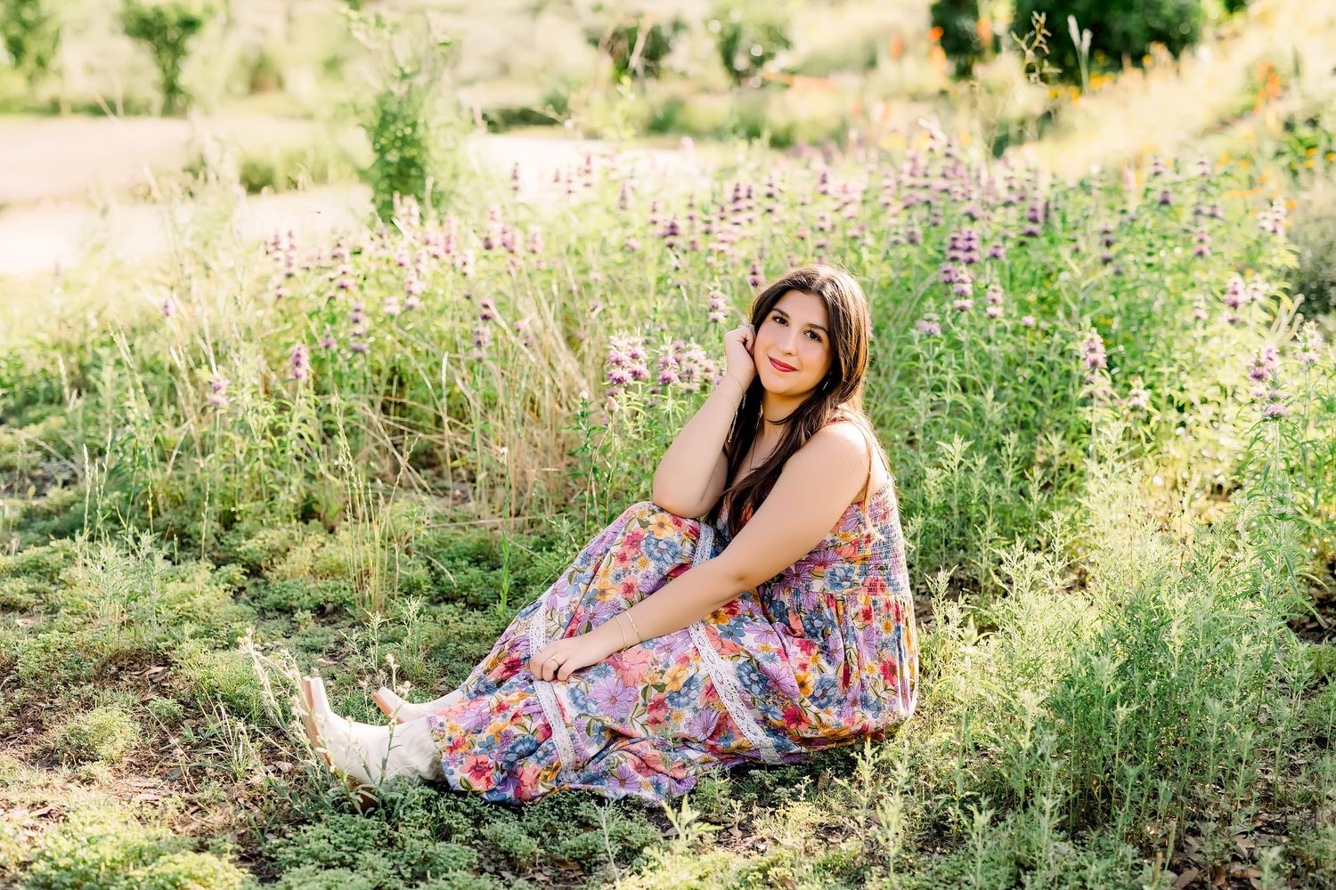 A brunette woman in a vibrant floral dress sits gracefully among wildflowers in a sunlit field.