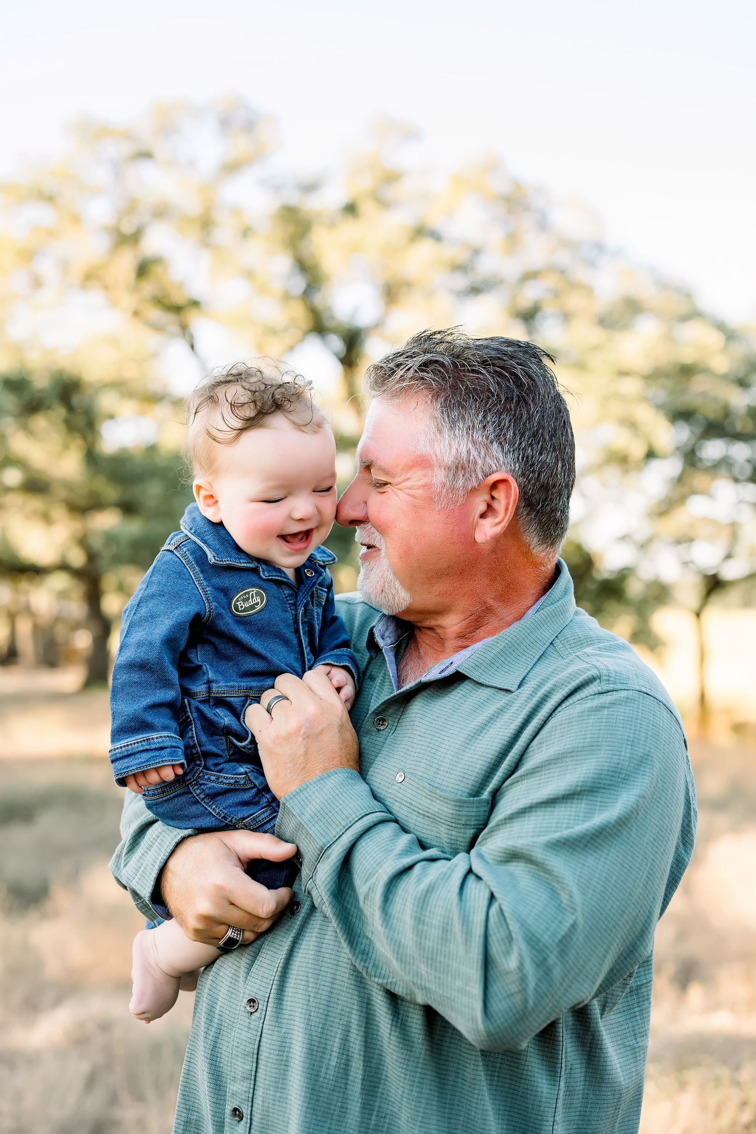 An older man with gray hair joyfully holds a laughing baby in a denim jacket outdoors in a sunlit field.