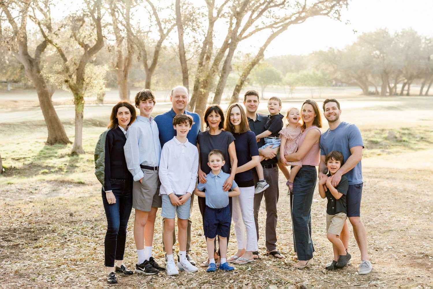 Multi-generational family portrait outdoors among trees, smiling together in warm golden light.