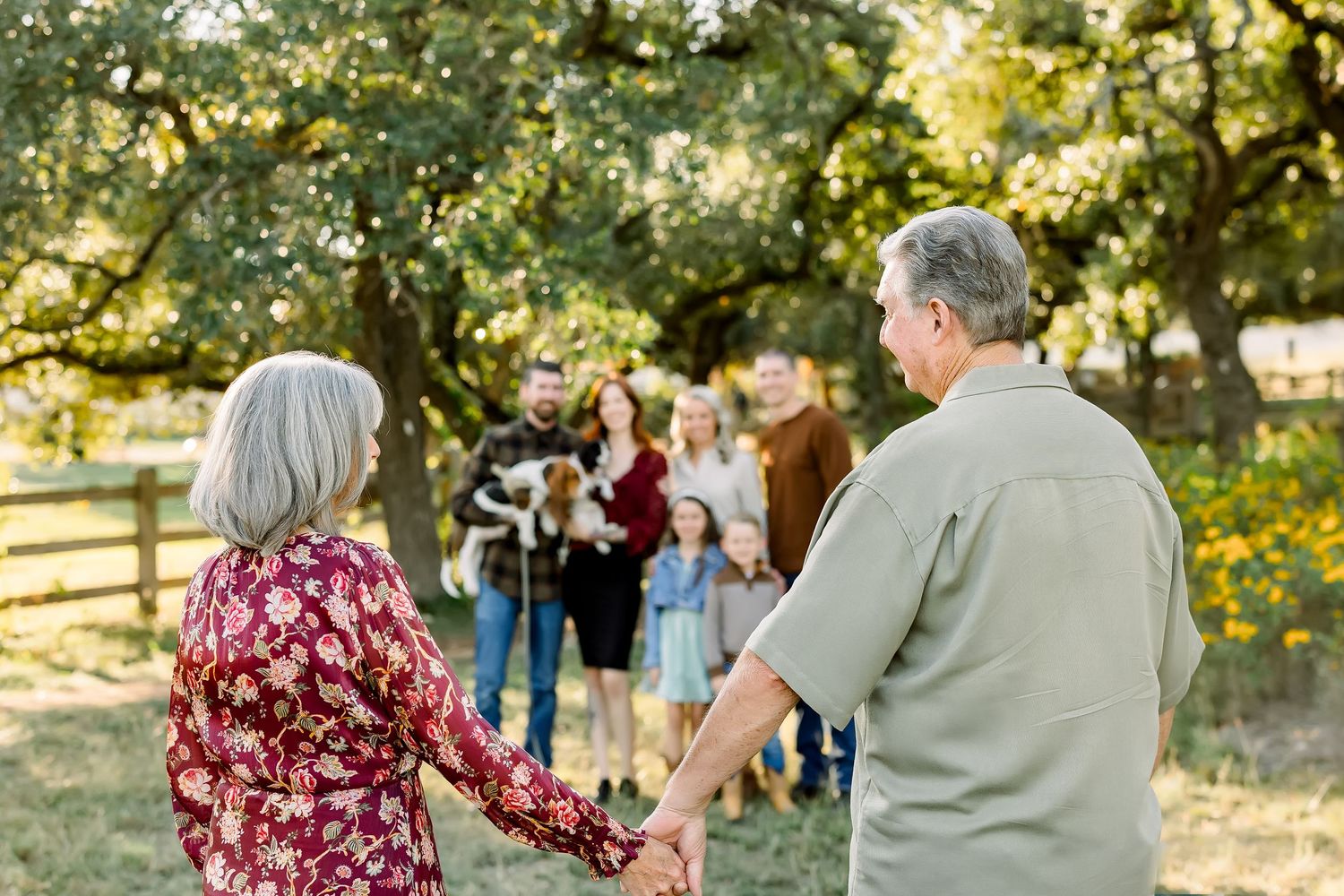 Older couple holding hands facing their multi-generational family gathered under oak trees during outdoor photo session.