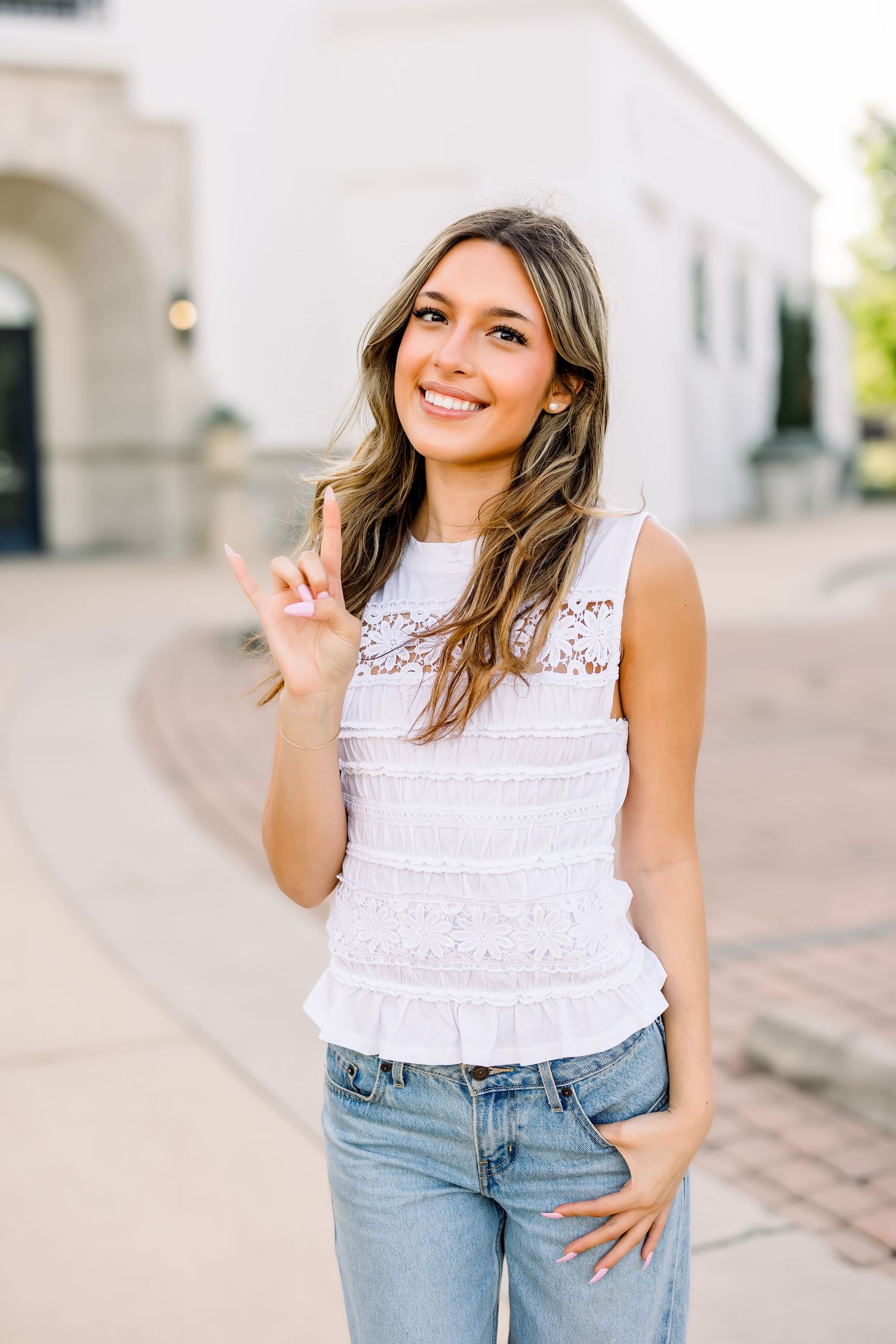 Smiling young woman in white lace sleeveless top and jeans making peace sign outdoors.