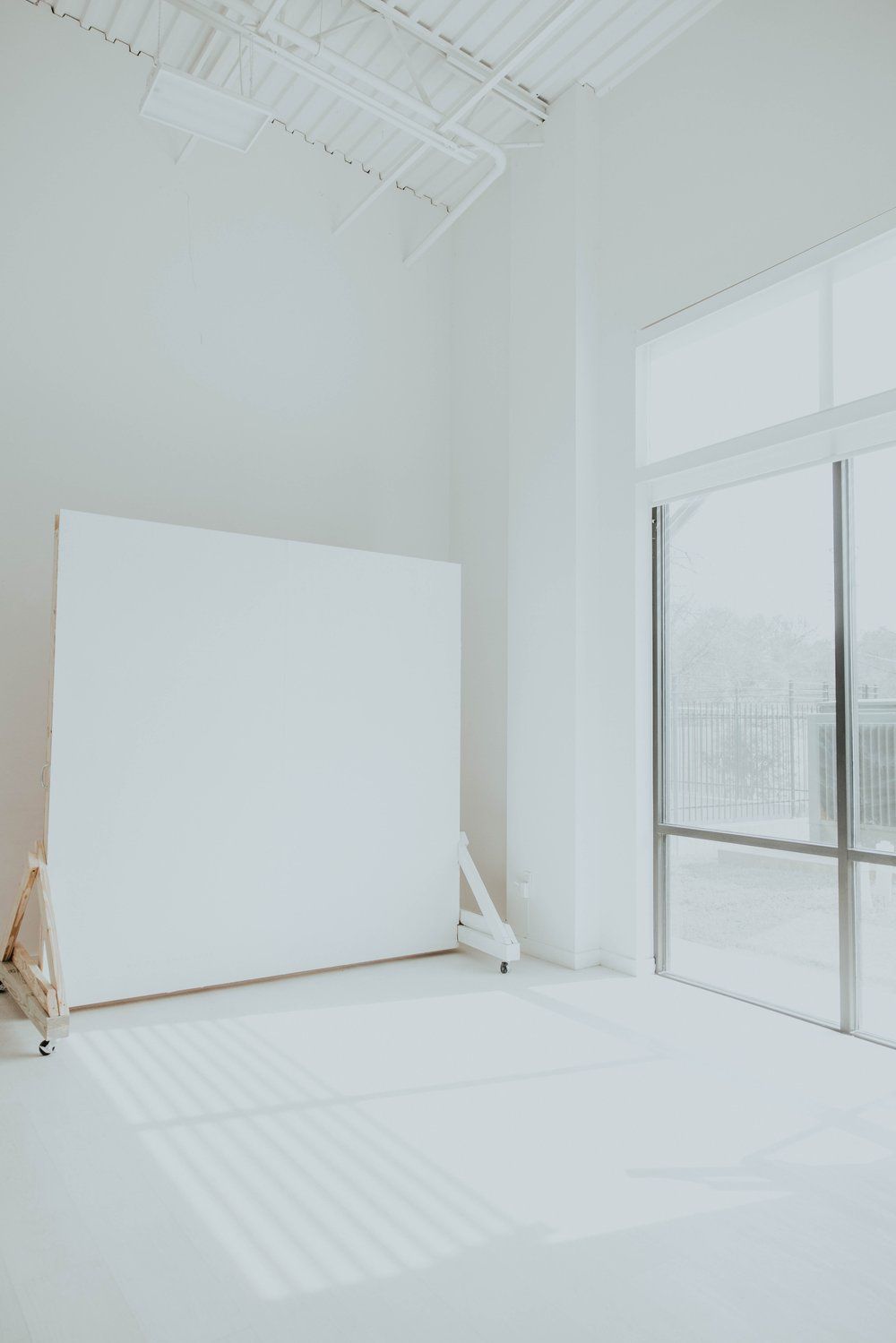 Bright white photography studio with large window, white backdrop on easel, and natural light casting soft shadows.