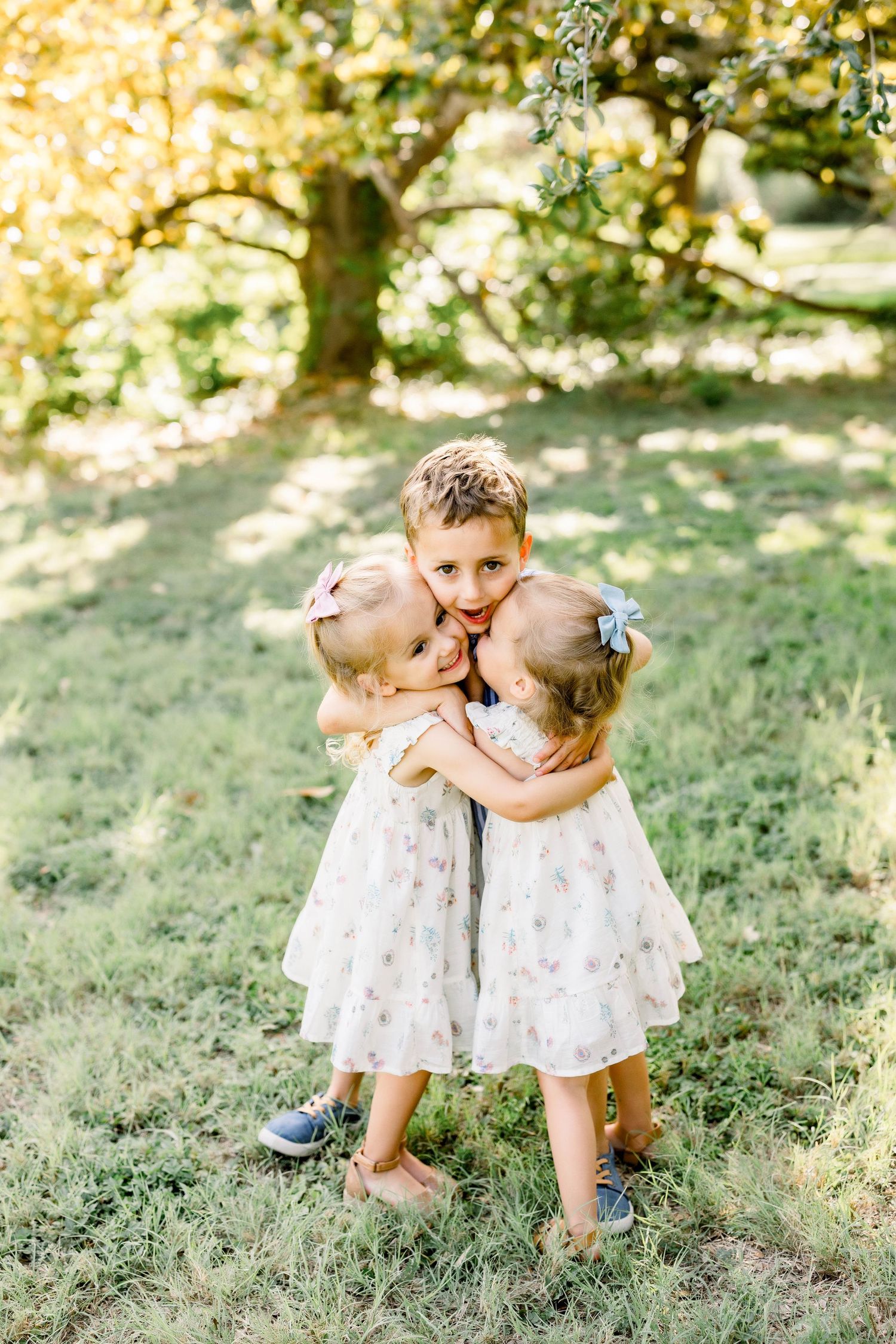 Three young siblings hugging outdoors under a tree, two girls in white floral dresses and a boy in the middle.