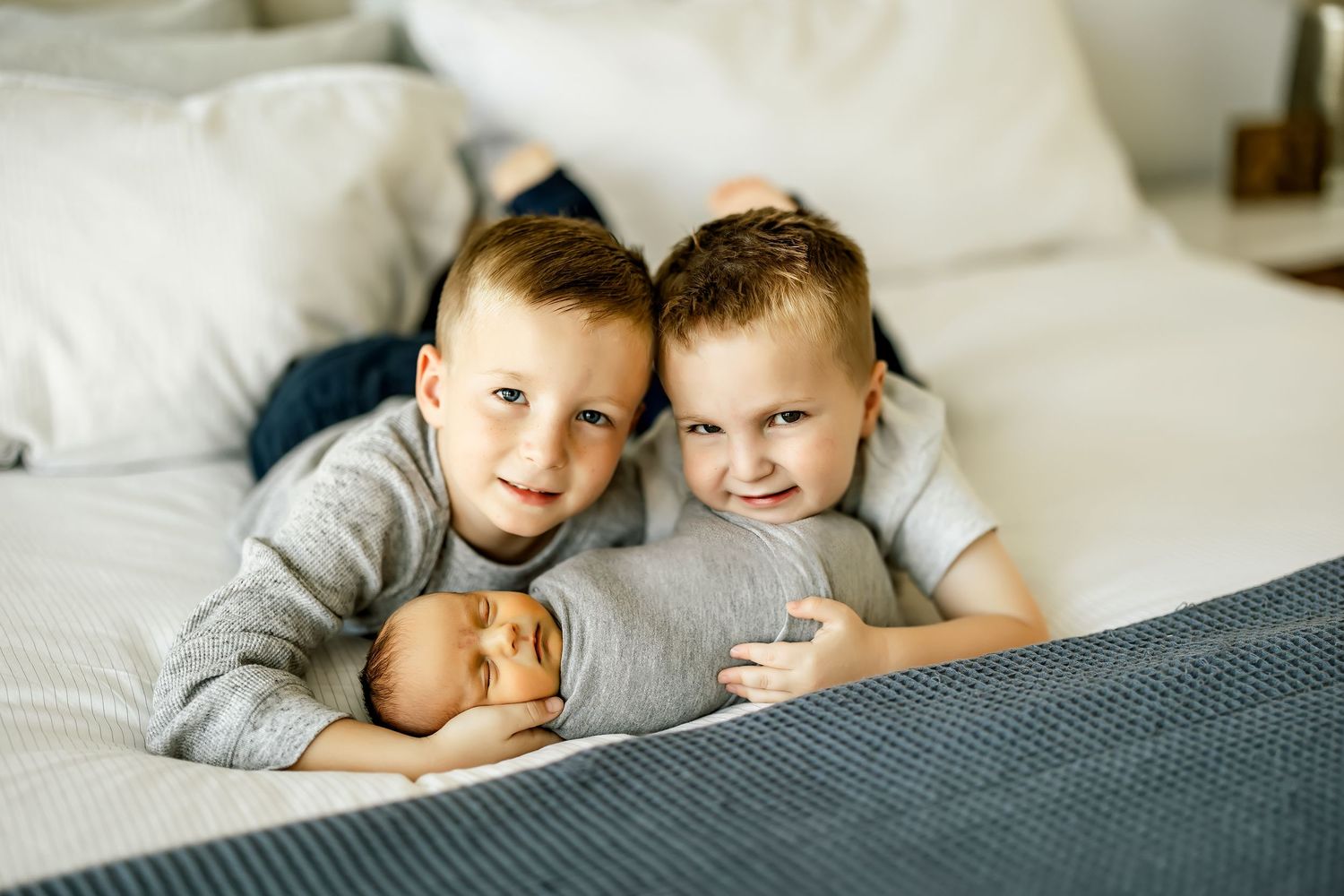Two young brothers in gray shirts smile while lying on a bed with their newborn baby sibling.