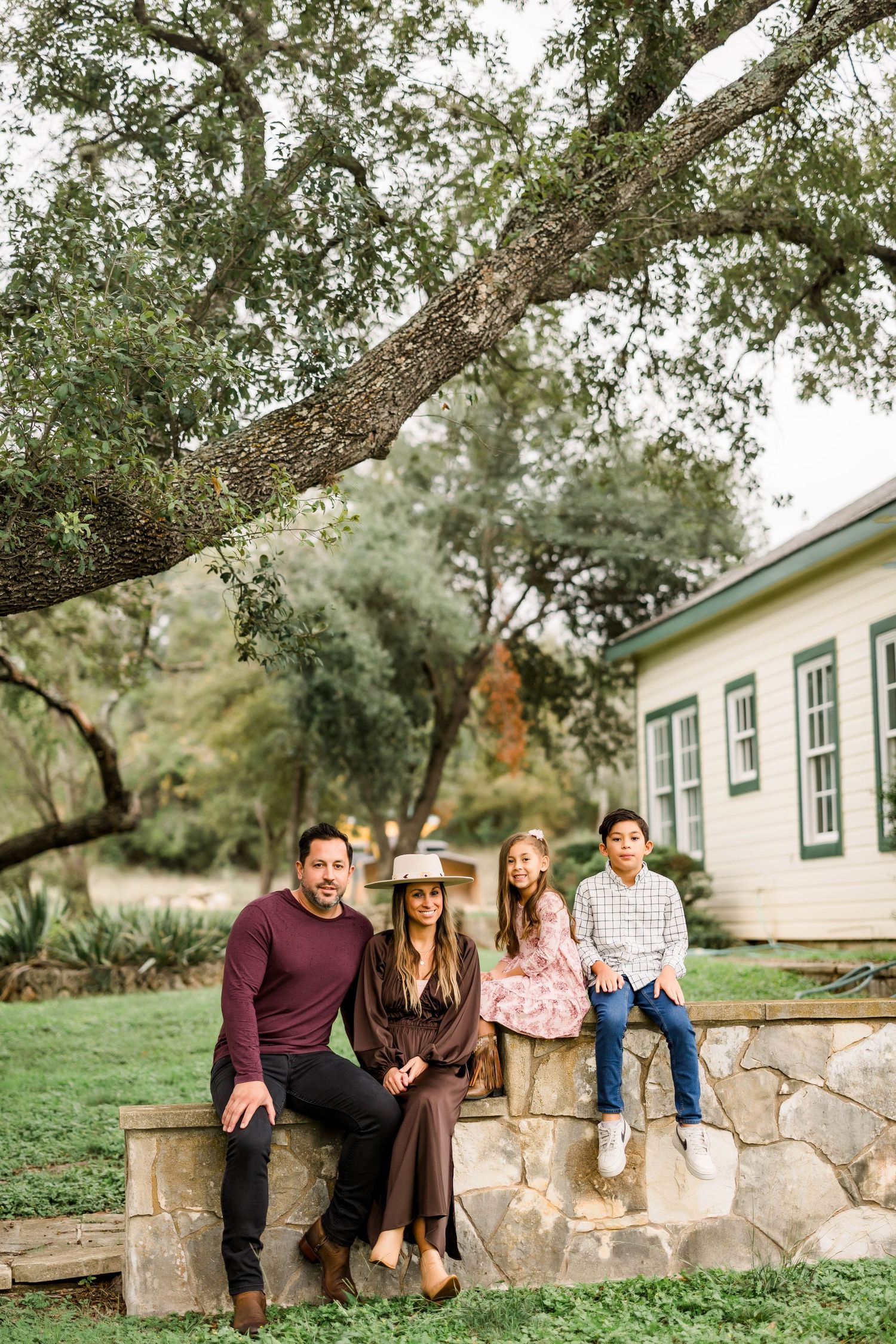 Family of four posing outdoors on a stone wall beneath a large oak tree near a charming country home.