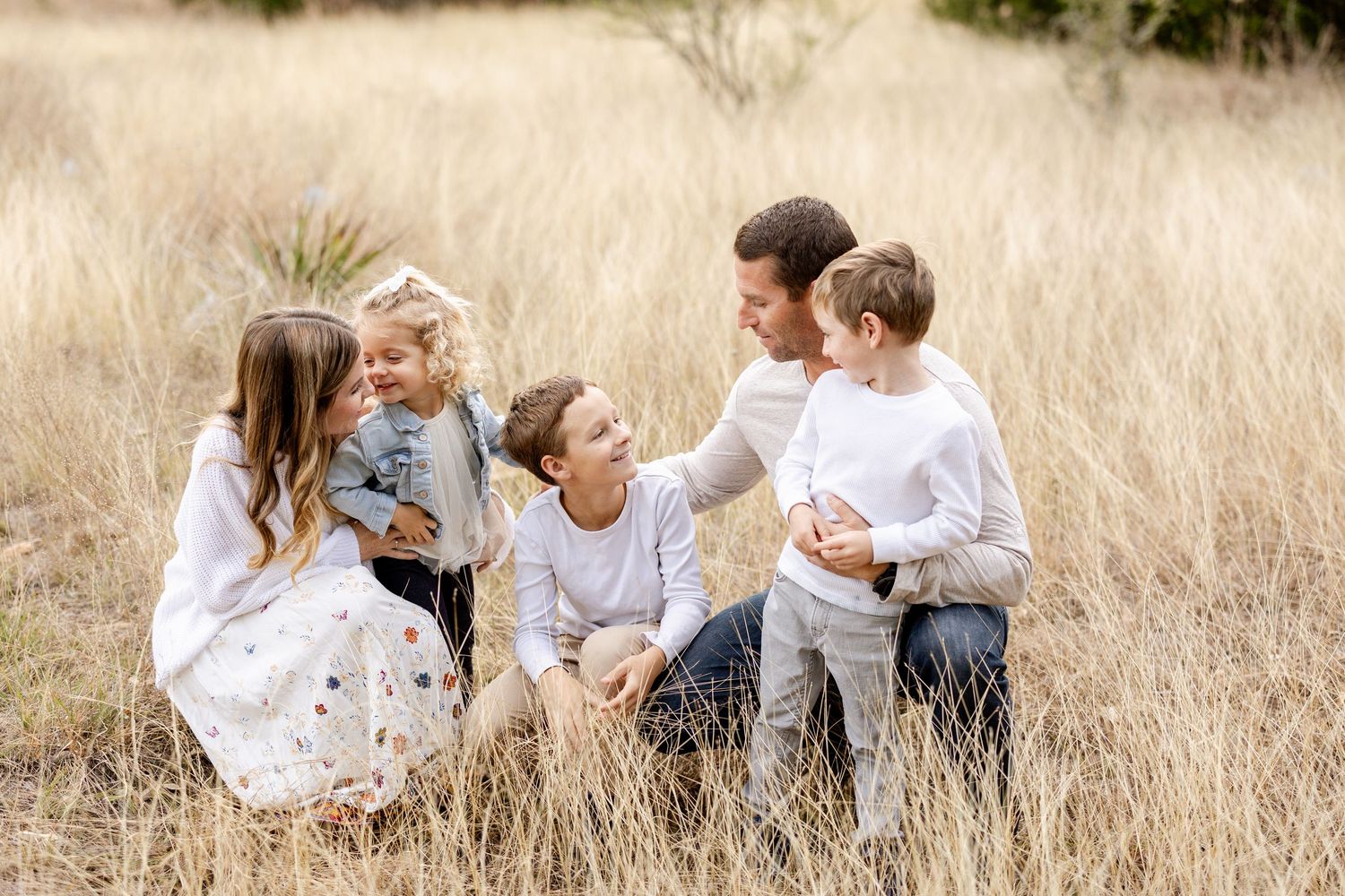 A joyful family of five shares a candid moment sitting together in tall golden grass during an outdoor portrait session.
