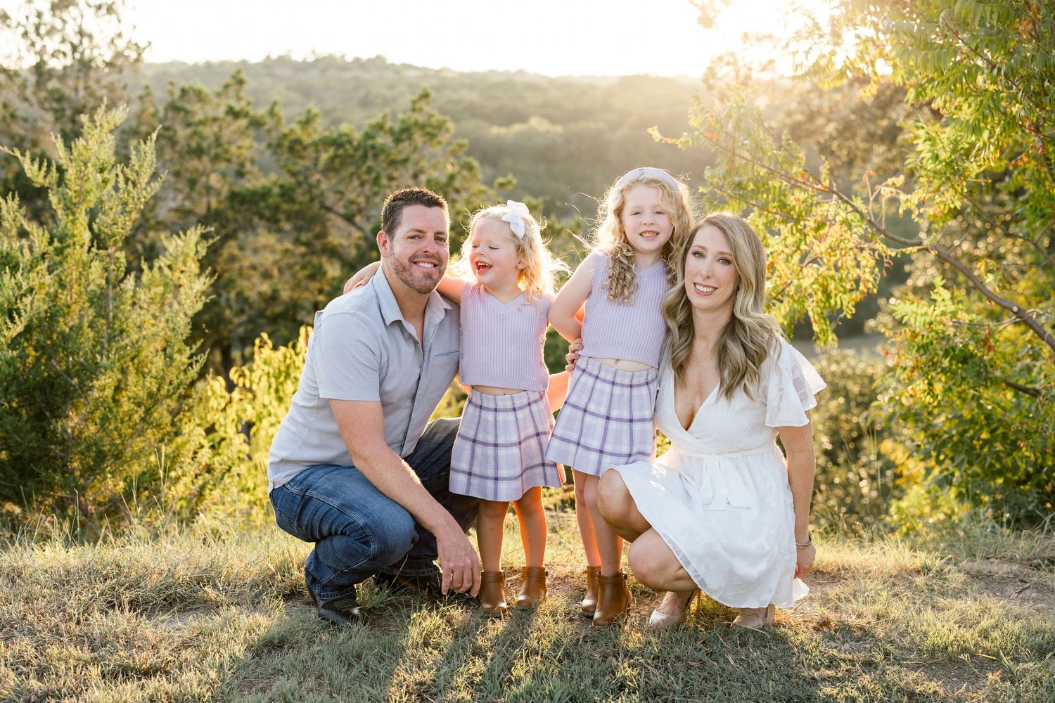 Smiling family of four posing outdoors on a grassy hill with warm golden sunset light in background.