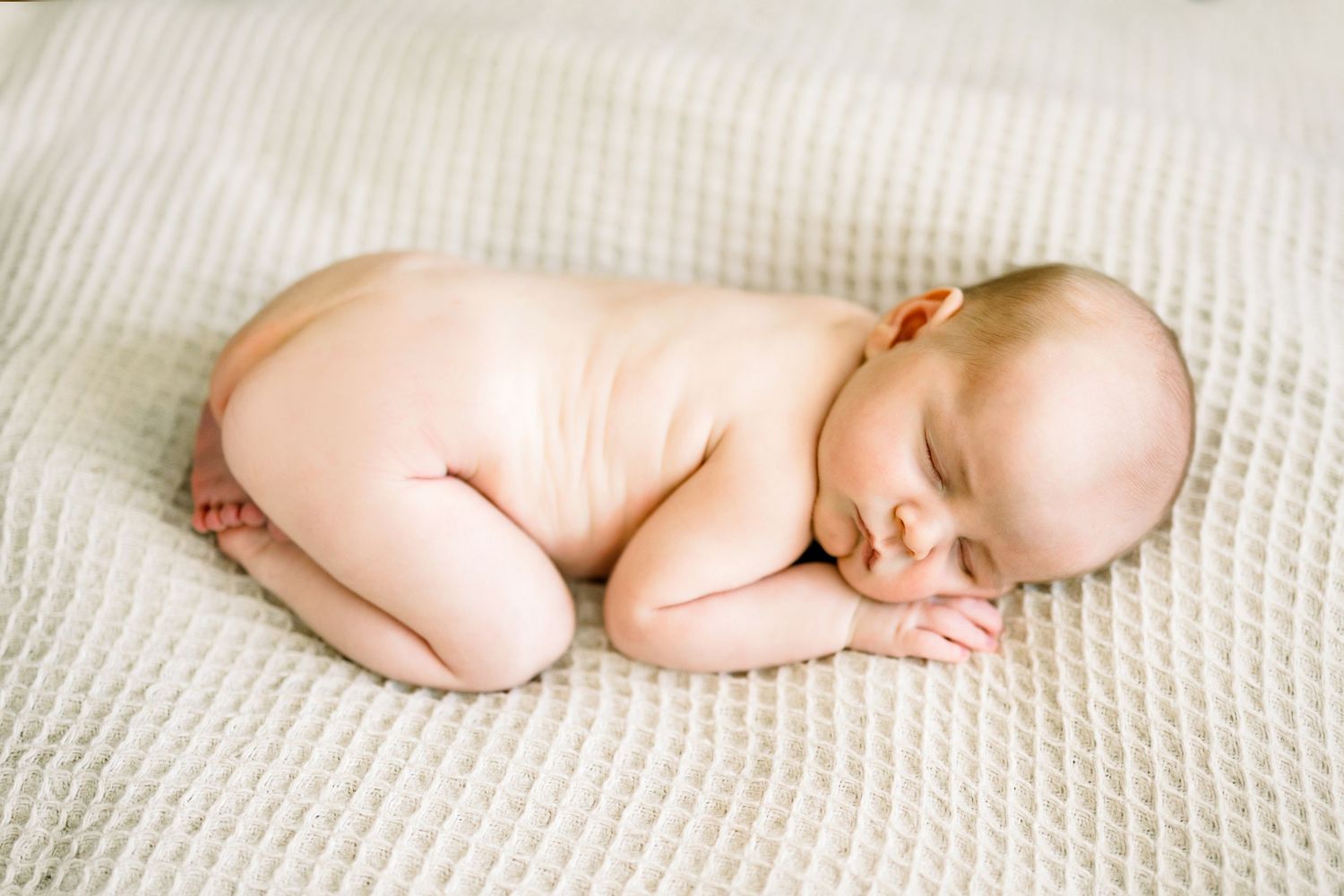 Peaceful naked newborn baby sleeping curled up on a soft cream textured blanket.