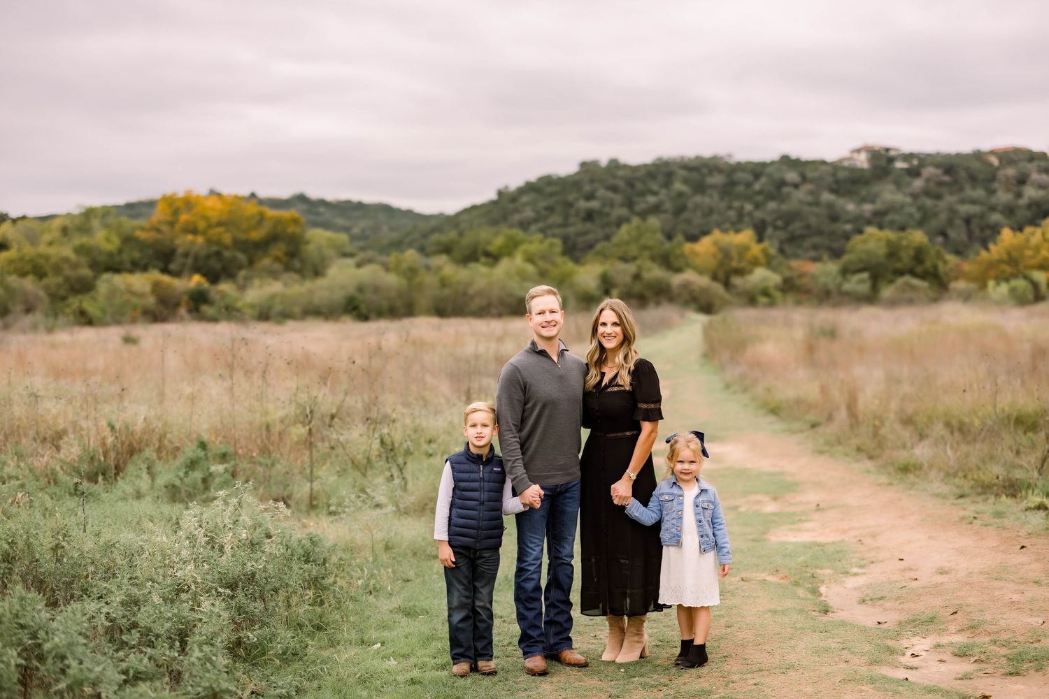 Family of four posing on a dirt path in an open field with rolling hills and autumn trees in the background.
