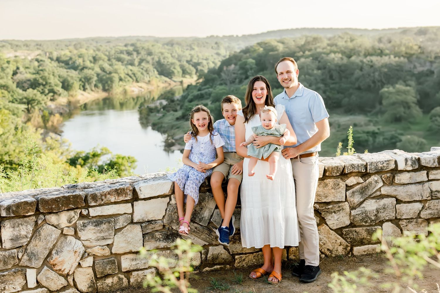 Happy family of five posing on stone wall overlooking scenic river and green landscape at golden hour.