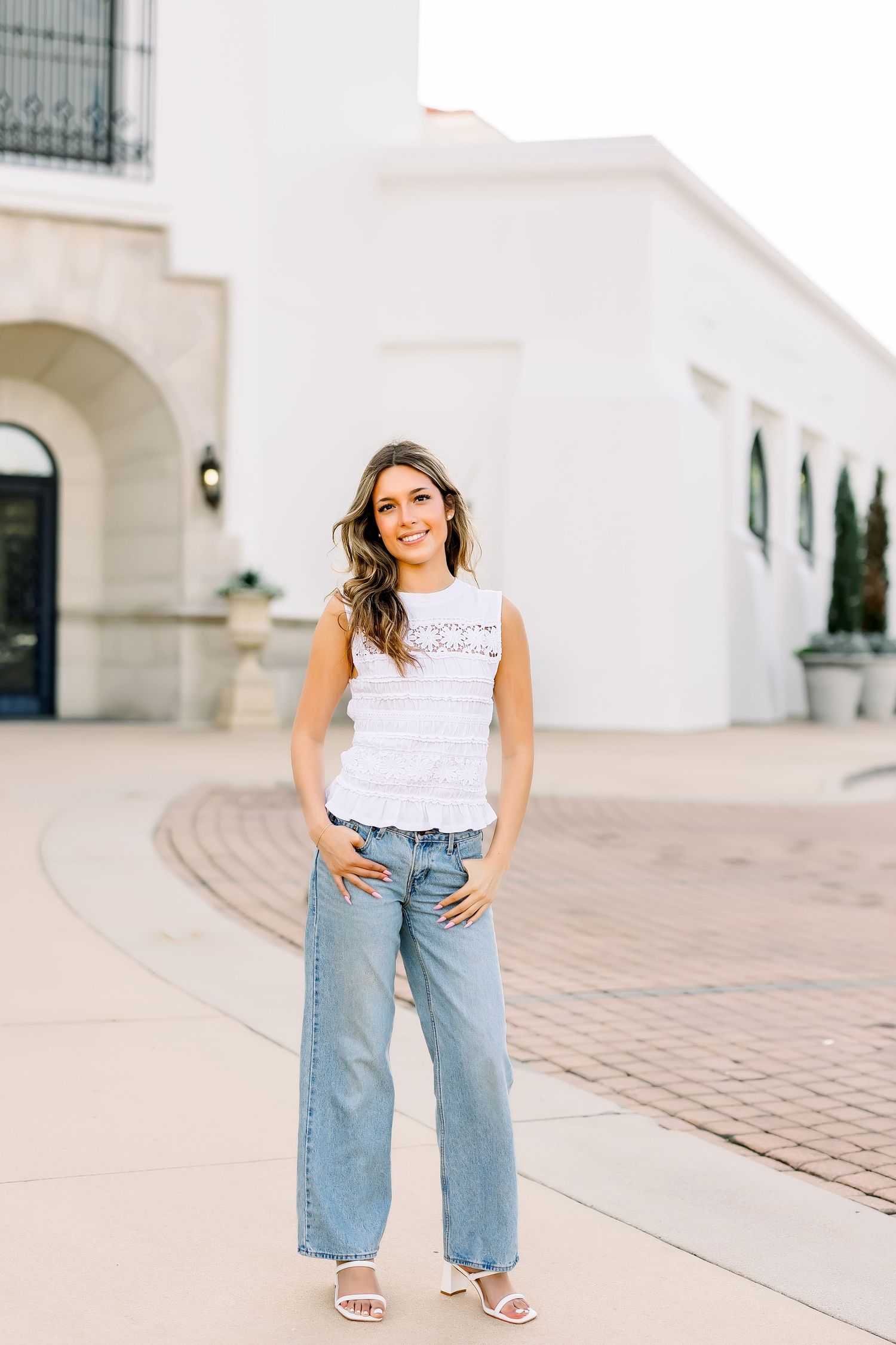 Young woman in white lace top and wide-leg jeans smiling with hands in pockets outside white building.