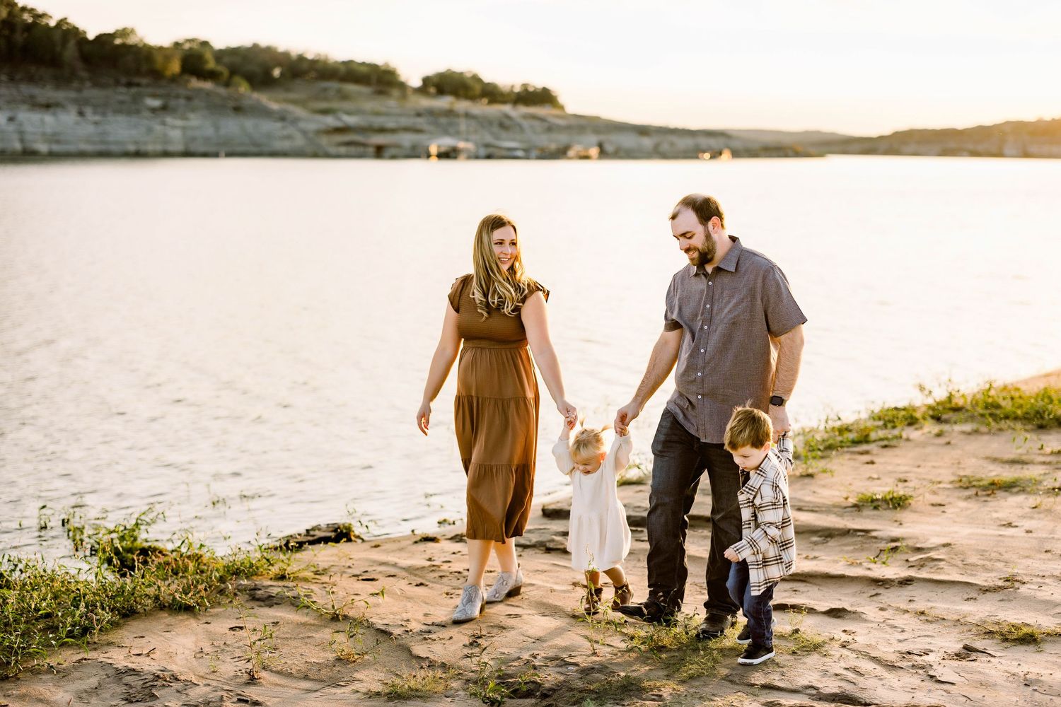 Family of four walking hand in hand along rocky lakeside at sunset.