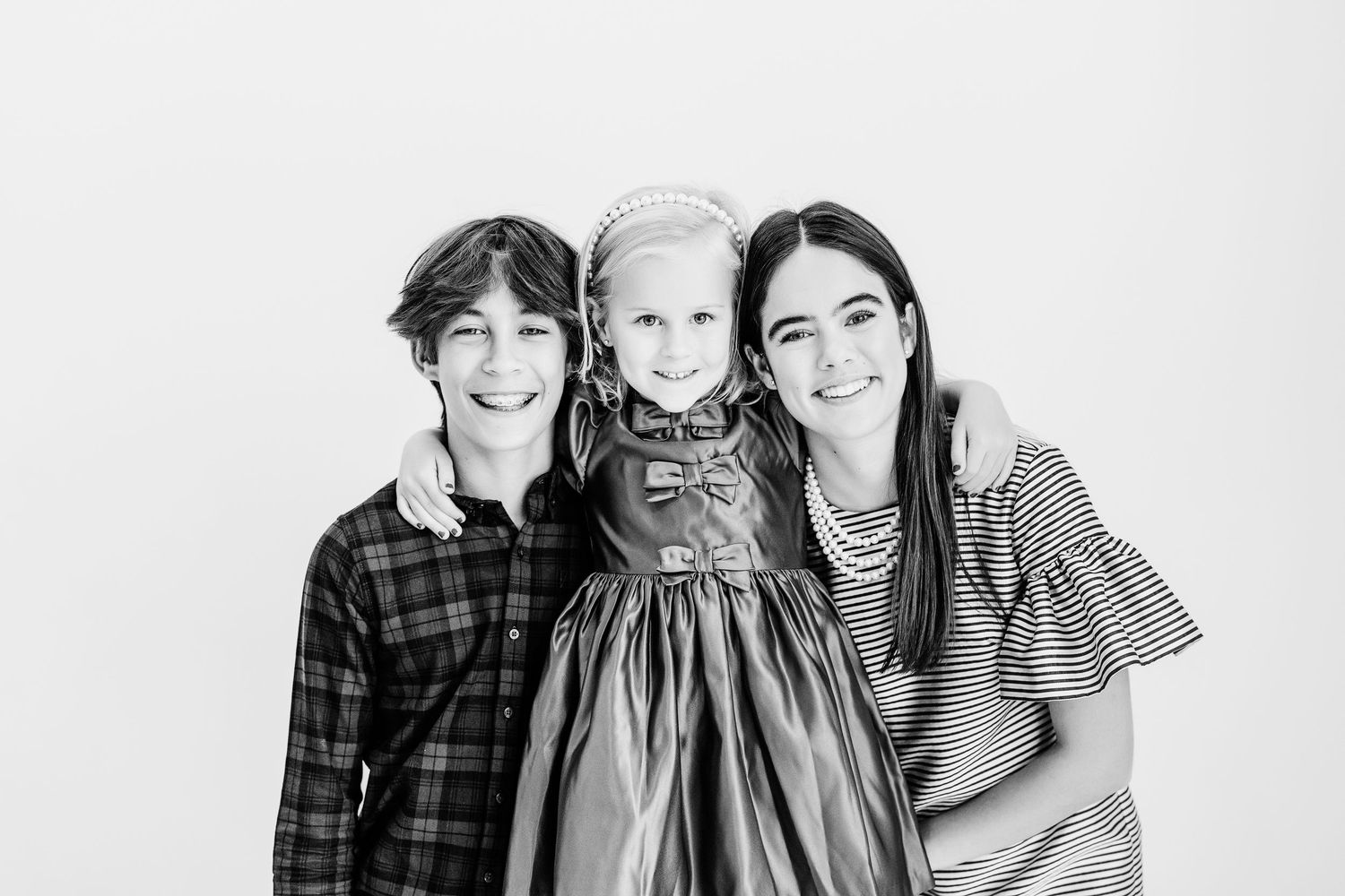 Black and white portrait of three siblings smiling together, dressed in plaid and striped outfits.