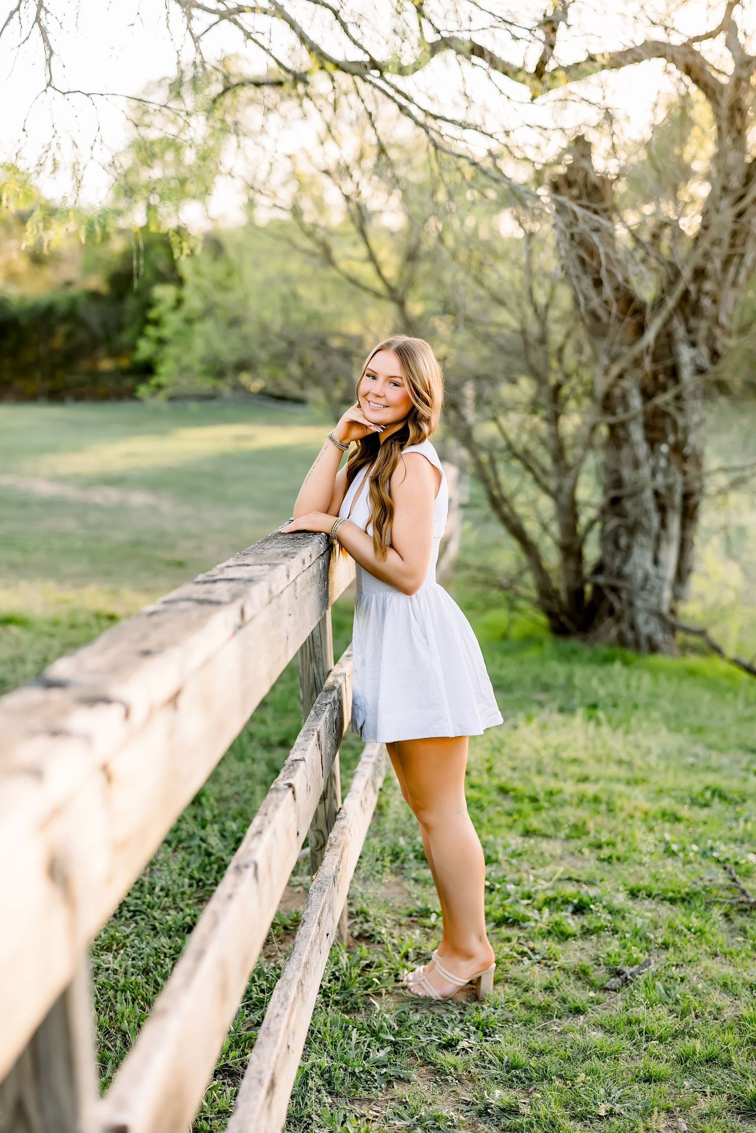 A smiling young woman in a white dress leans against a rustic wooden fence in a green meadow.