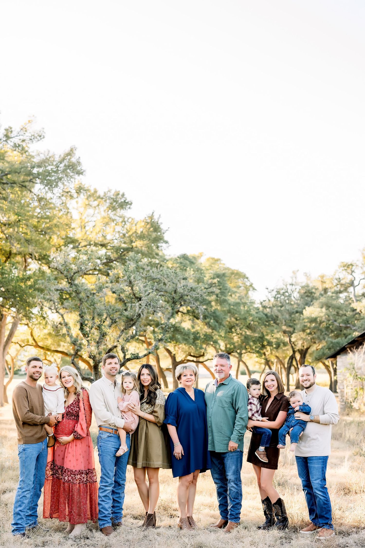 Large family posing outdoors in coordinated fall outfits among golden oak trees during a professional portrait session.