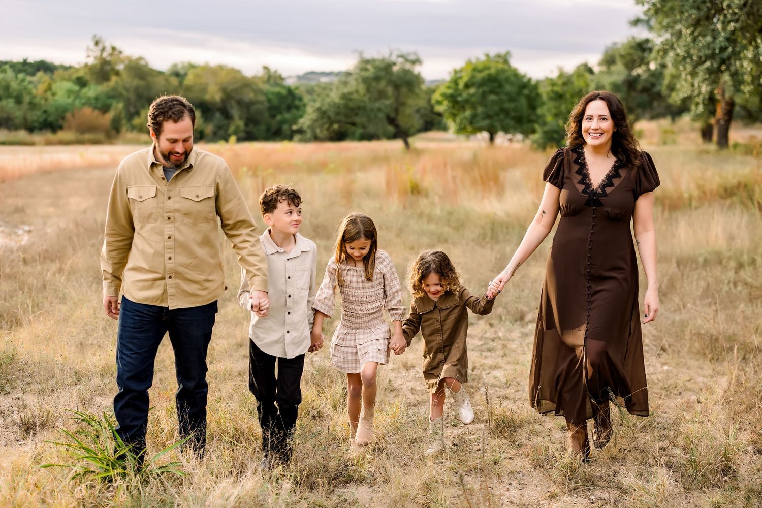 Family of five walks hand in hand through golden meadow with lush green trees behind them.