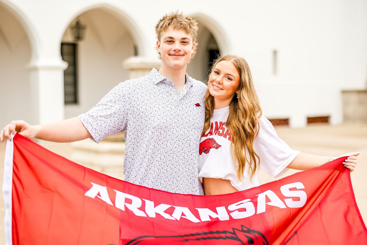 Young couple smiling and holding red Arkansas Razorbacks flag outside white building.