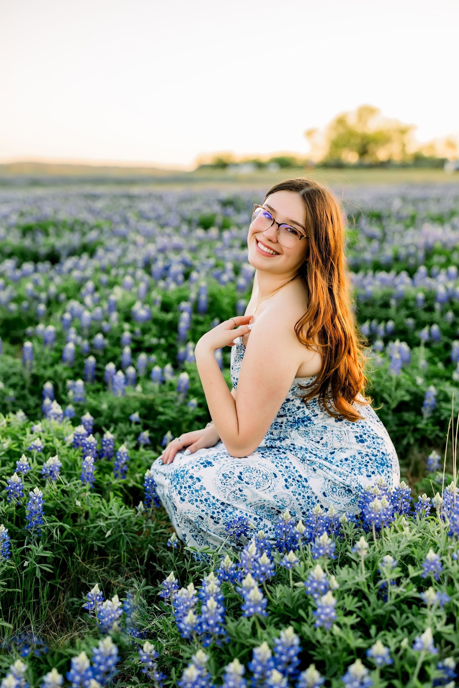 Smiling teenage girl in a floral dress sitting in a field of Texas bluebonnets at sunset.