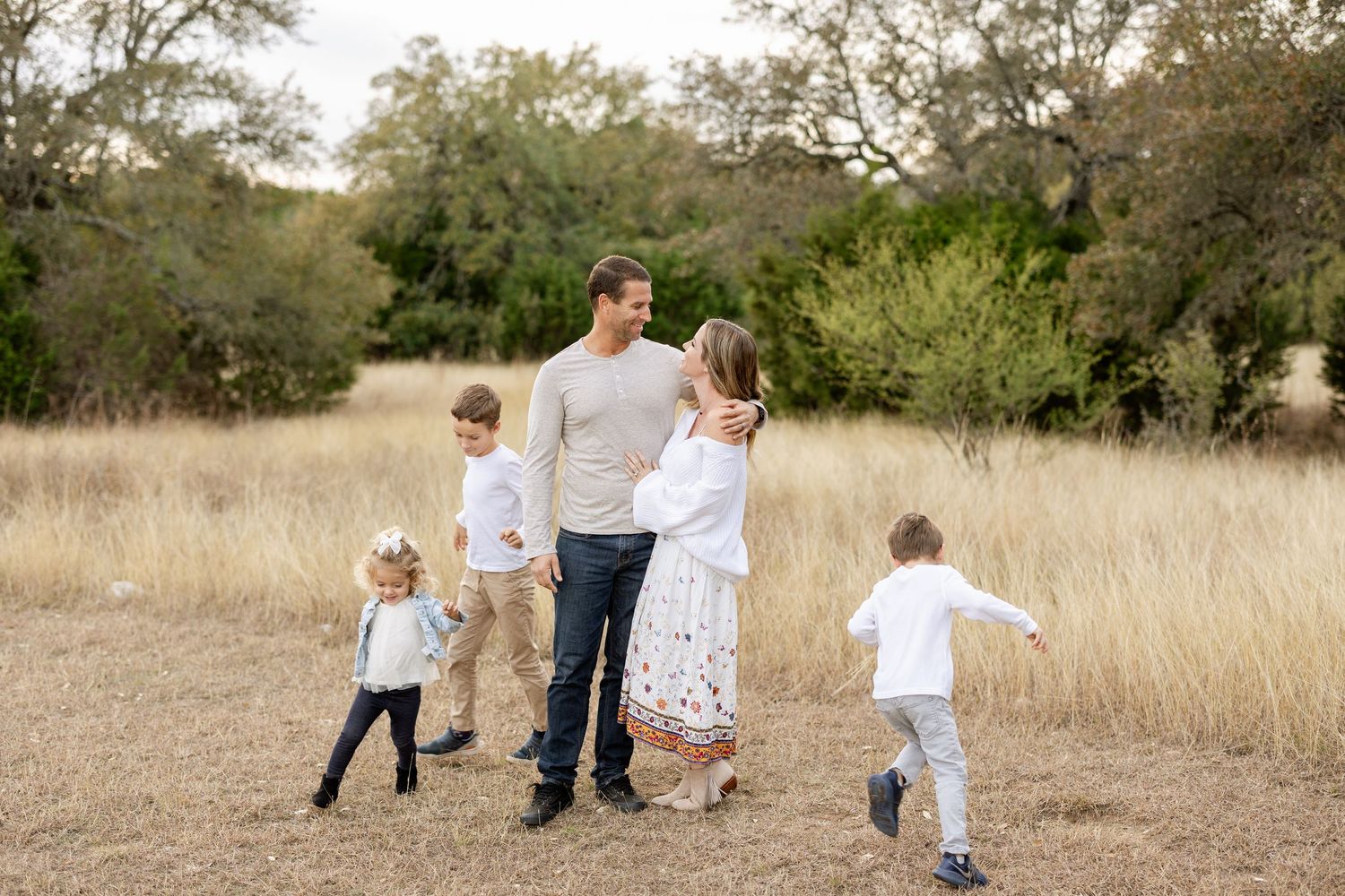 A family of five enjoys a candid walk through a golden meadow surrounded by oak trees during an outdoor photo session.