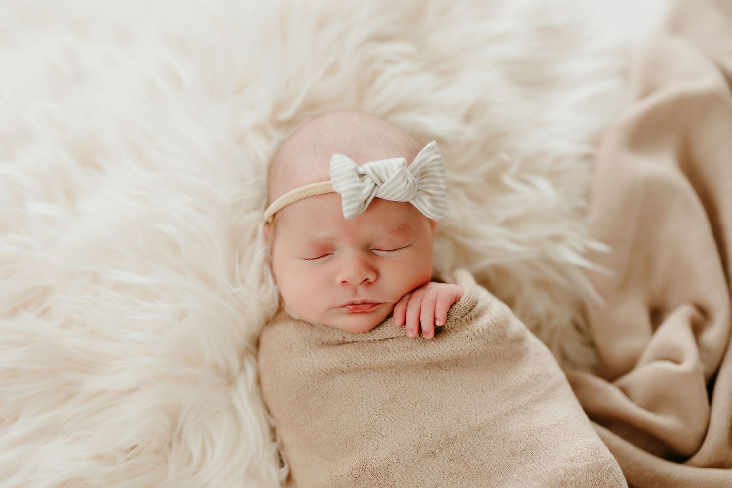 Sleeping newborn baby girl swaddled in beige wrap with a bow headband on a fluffy white fur blanket.