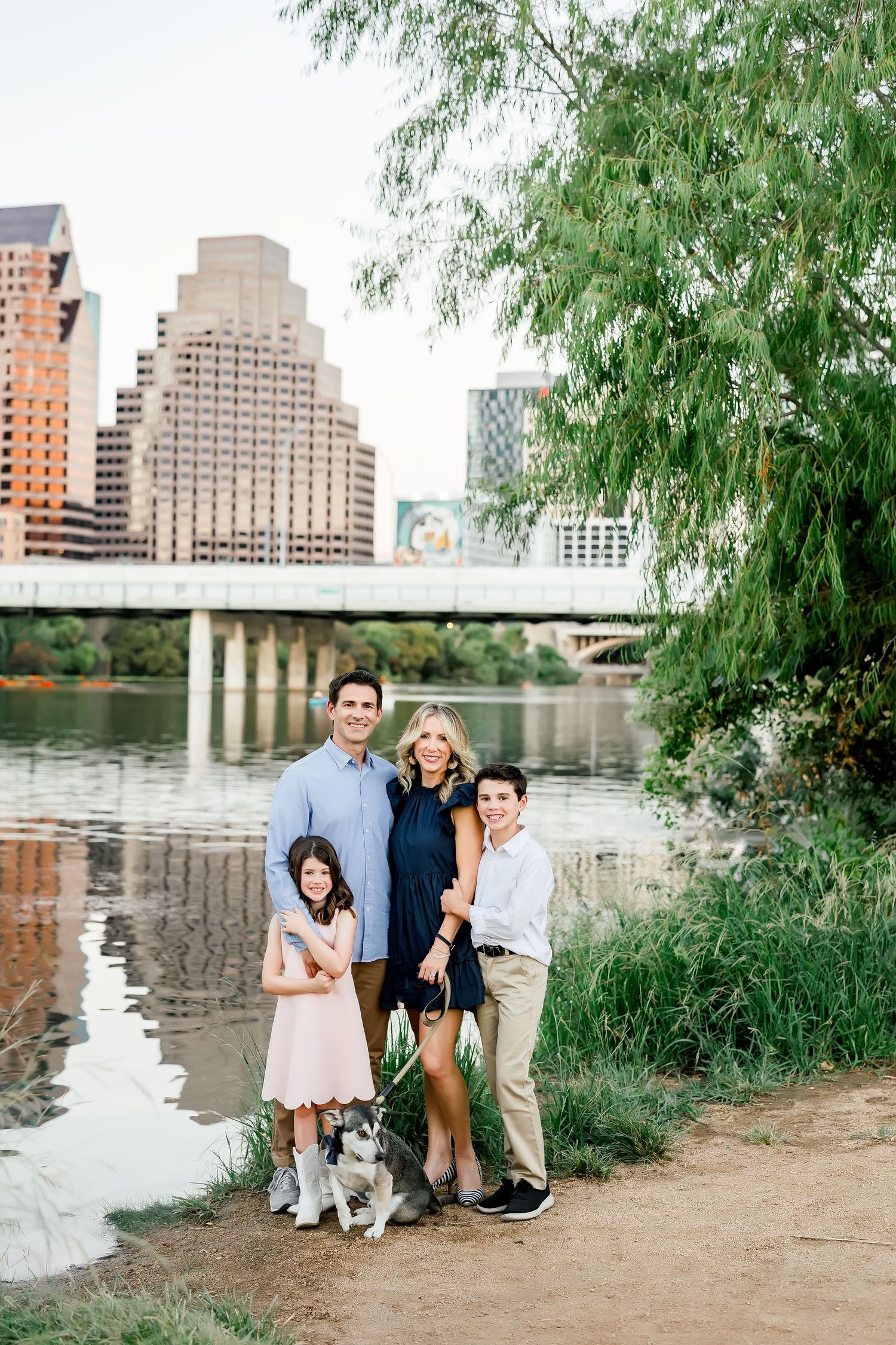 Family of four with a husky dog poses along a riverbank with the Austin, TX city skyline behind them.