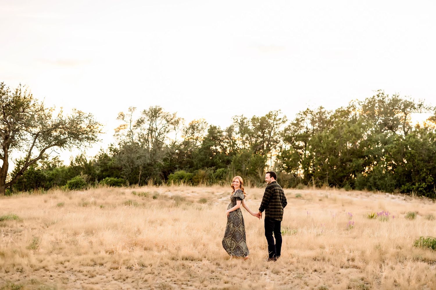 A couple holds hands while walking through a golden field at sunset with trees in the background.