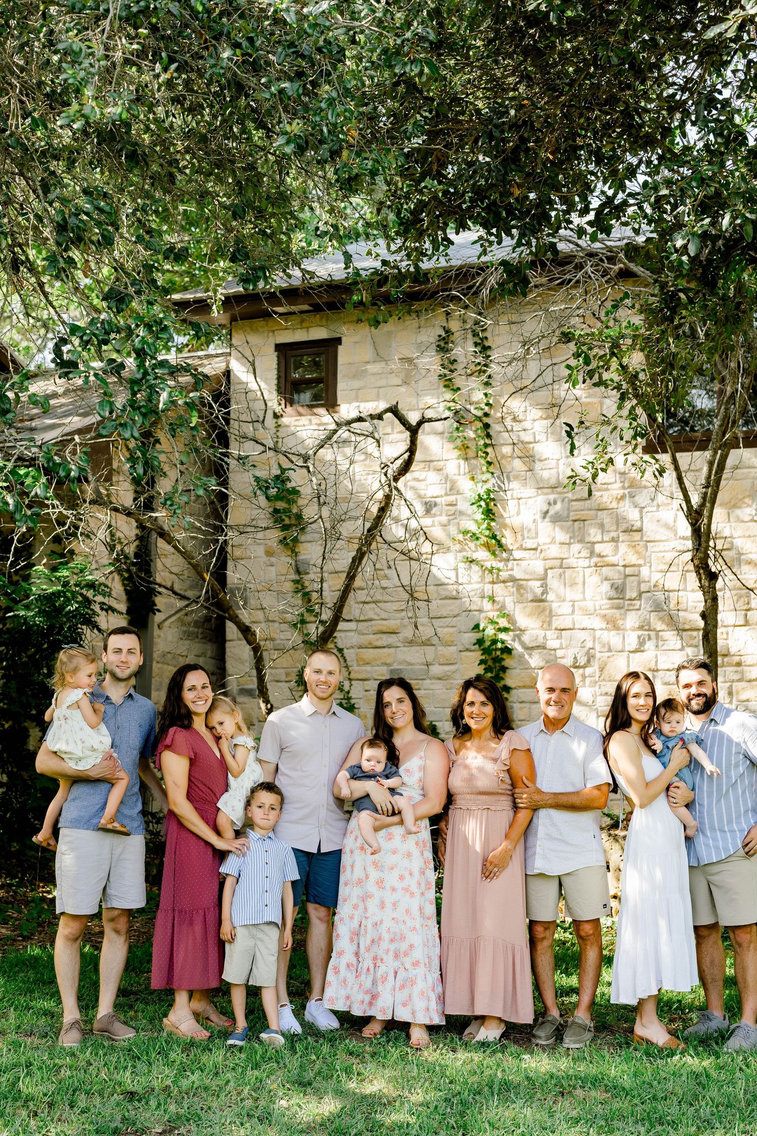 Large family group portrait outdoors on green lawn with stone building and lush trees, including adults, children, and a newborn baby.