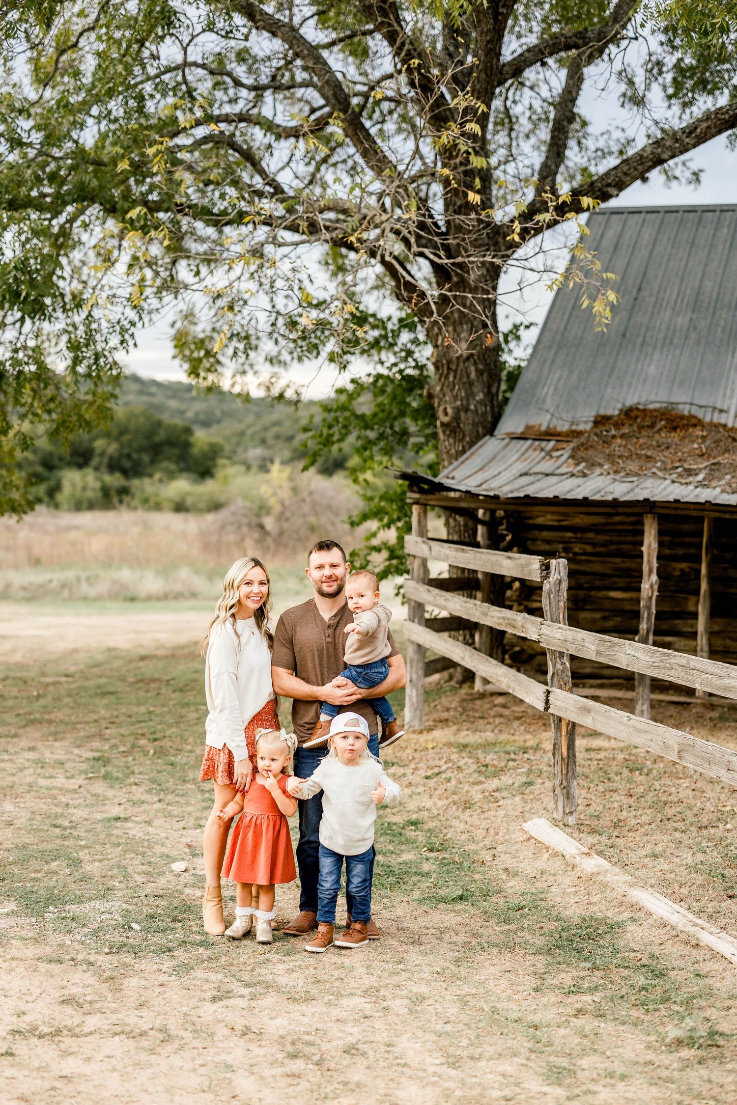 Family of five posing near a rustic barn and large oak tree outdoors at a mini session in Commons Ford Ranch in Bee Cave, Tx.