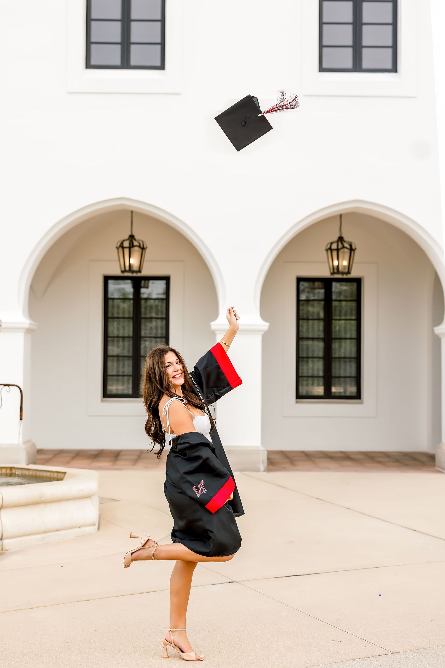 Young woman in graduation gown joyfully tossing cap in air outside white arched building.