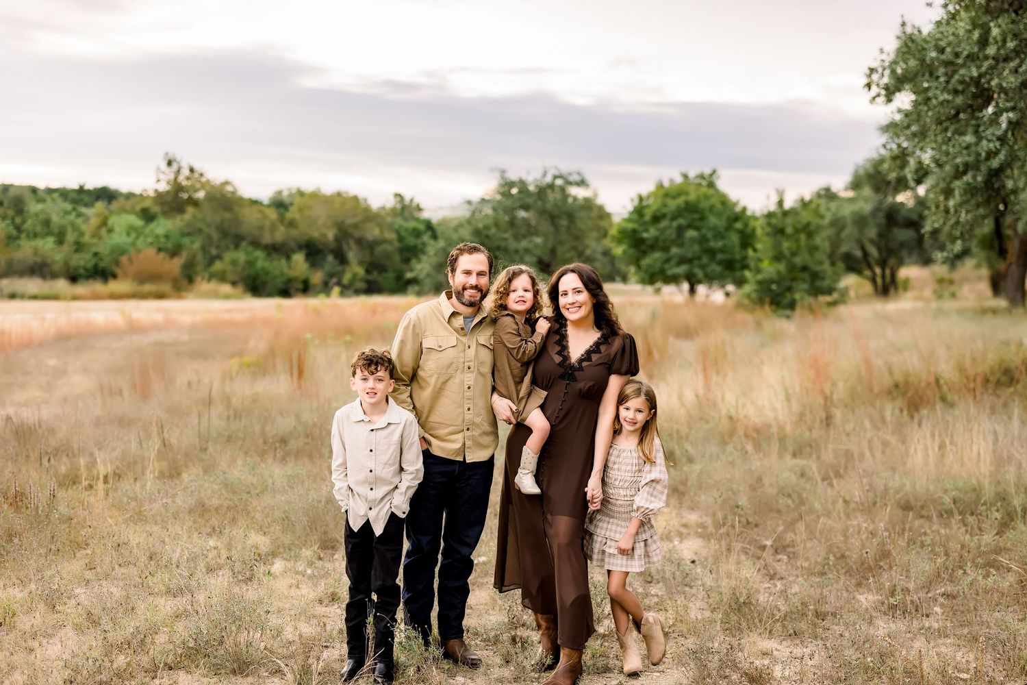 Family of five smiling in golden field at dusk, parents with three children in earth tones.