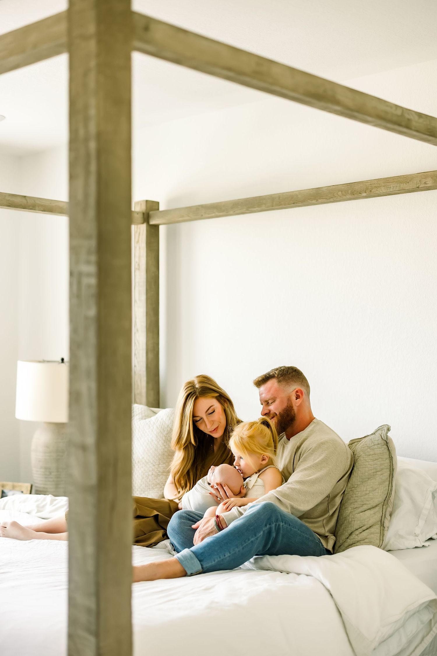 A couple sits on a four-poster bed with their toddler and newborn baby in a cozy white bedroom.