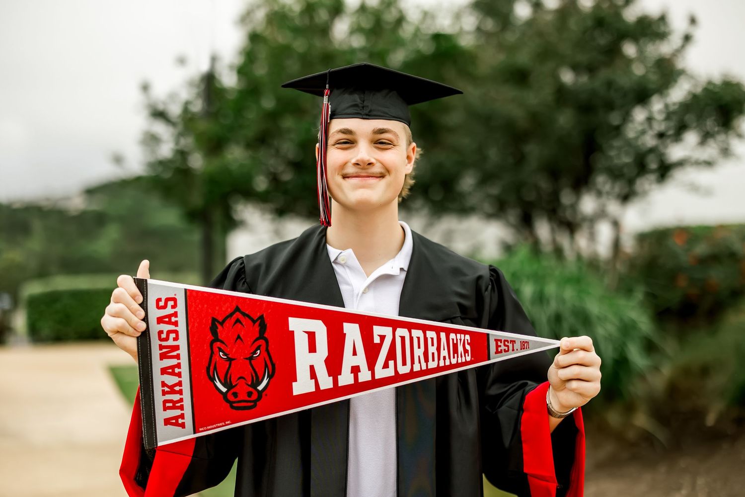 Smiling graduate in cap and gown holding Arkansas Razorbacks pennant outdoors.