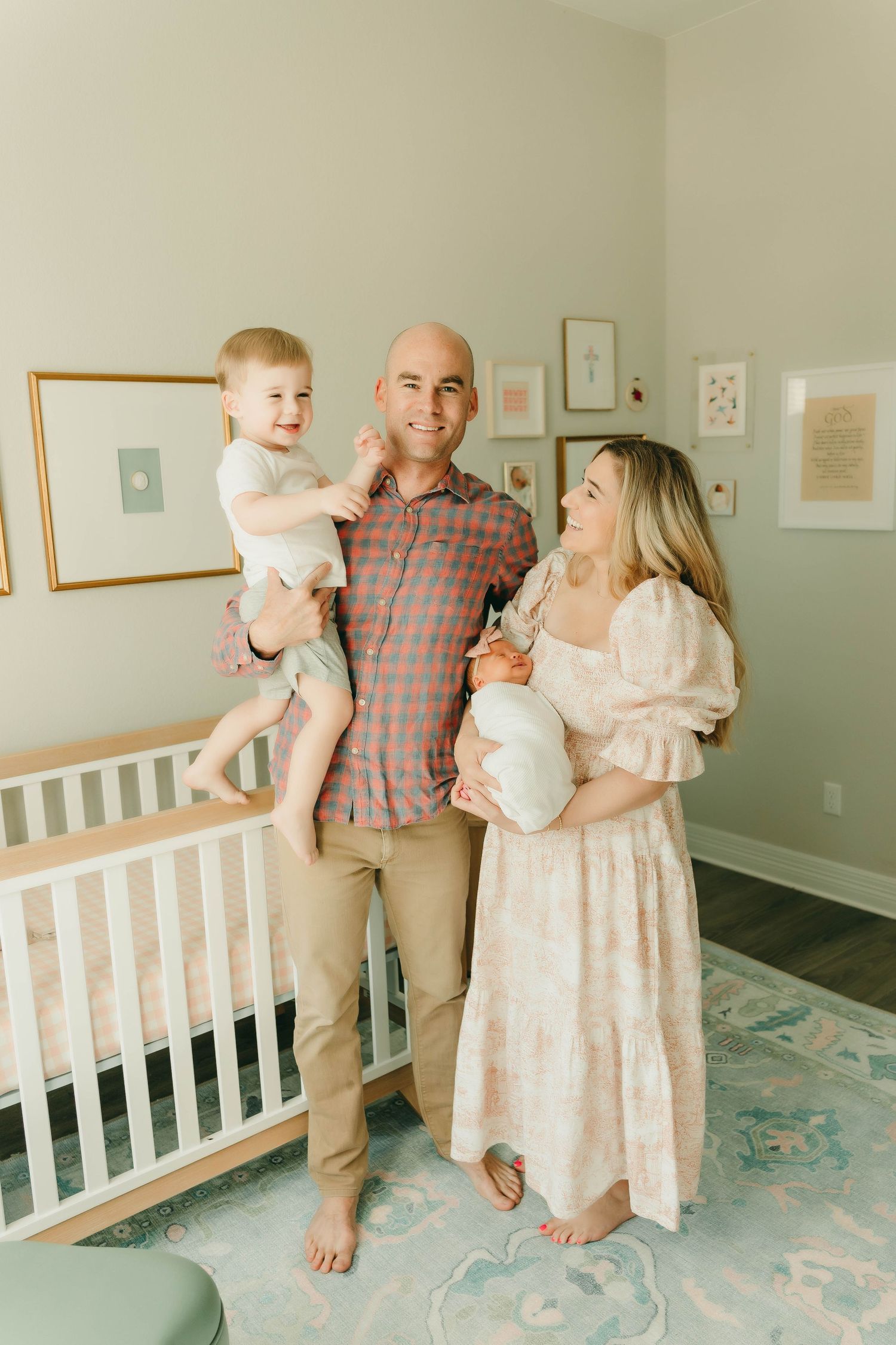 Family of four posing in a pastel nursery with parents holding their toddler and newborn.