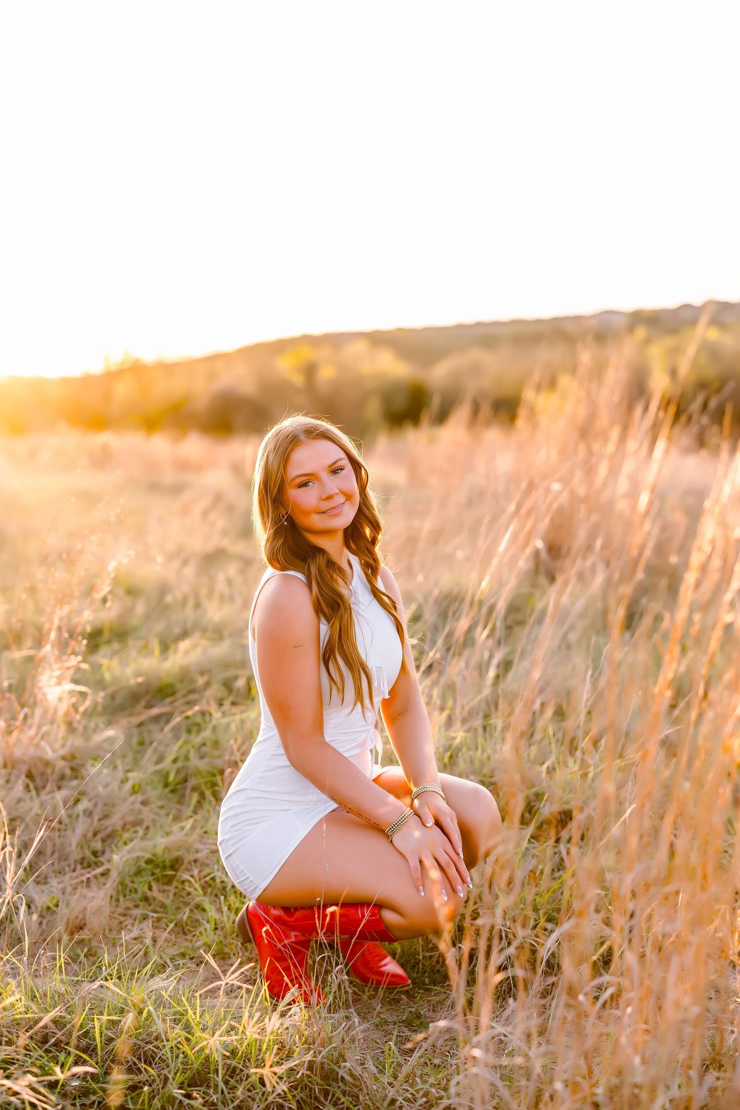 A smiling young woman in a white dress and red cowboy boots poses in a golden sunset field.