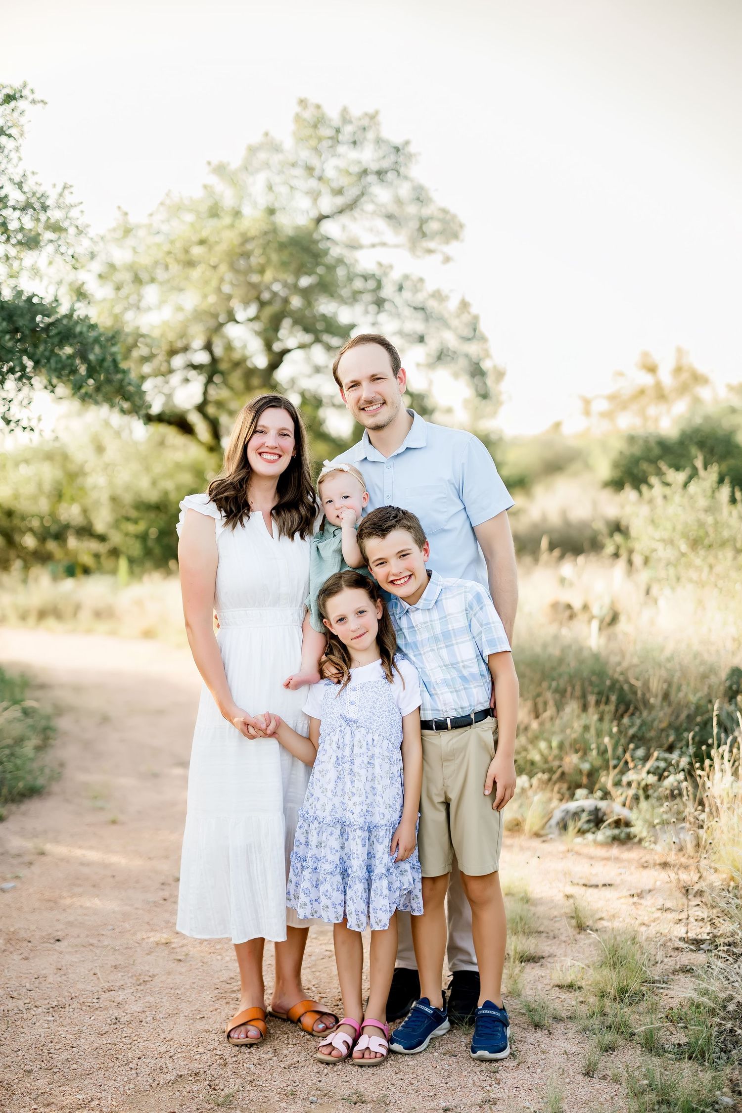 Family of five smiling on a sunny dirt path surrounded by lush greenery, parents with three young children.
