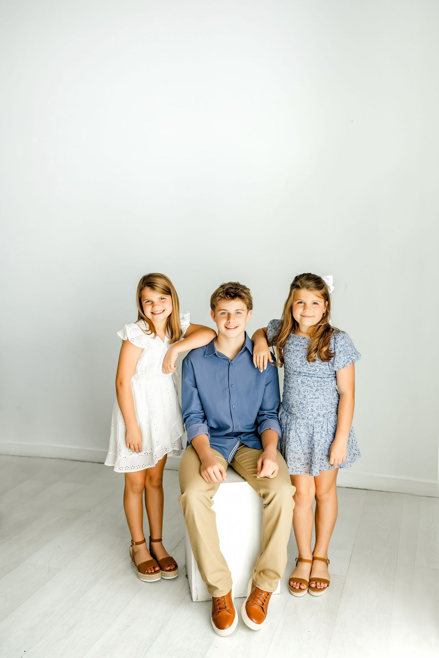 Three siblings posing together, boy in blue shirt seated between two girls in dresses against white wall.