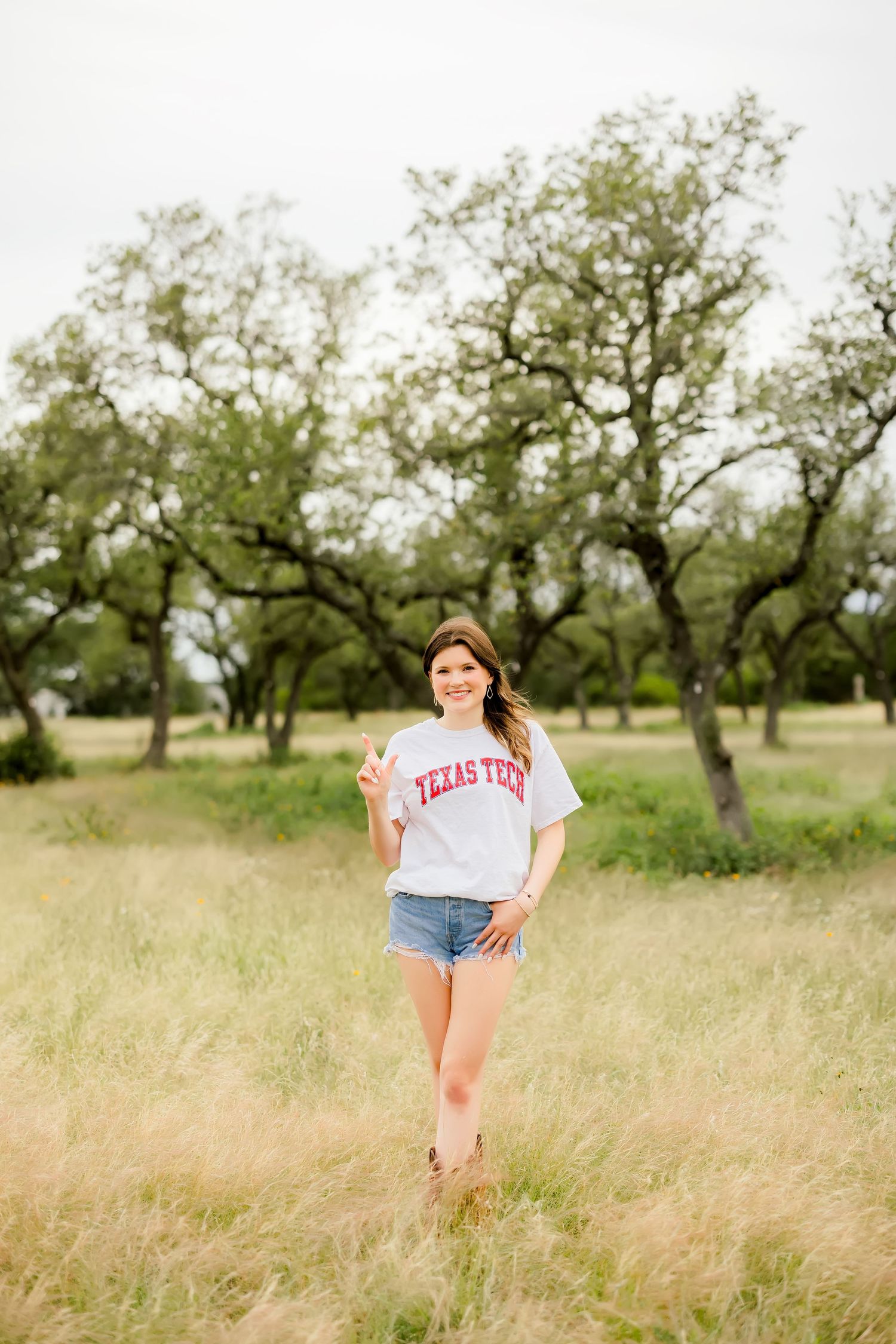 Young woman in Texas Tech shirt and denim shorts making peace sign in green meadow with trees.