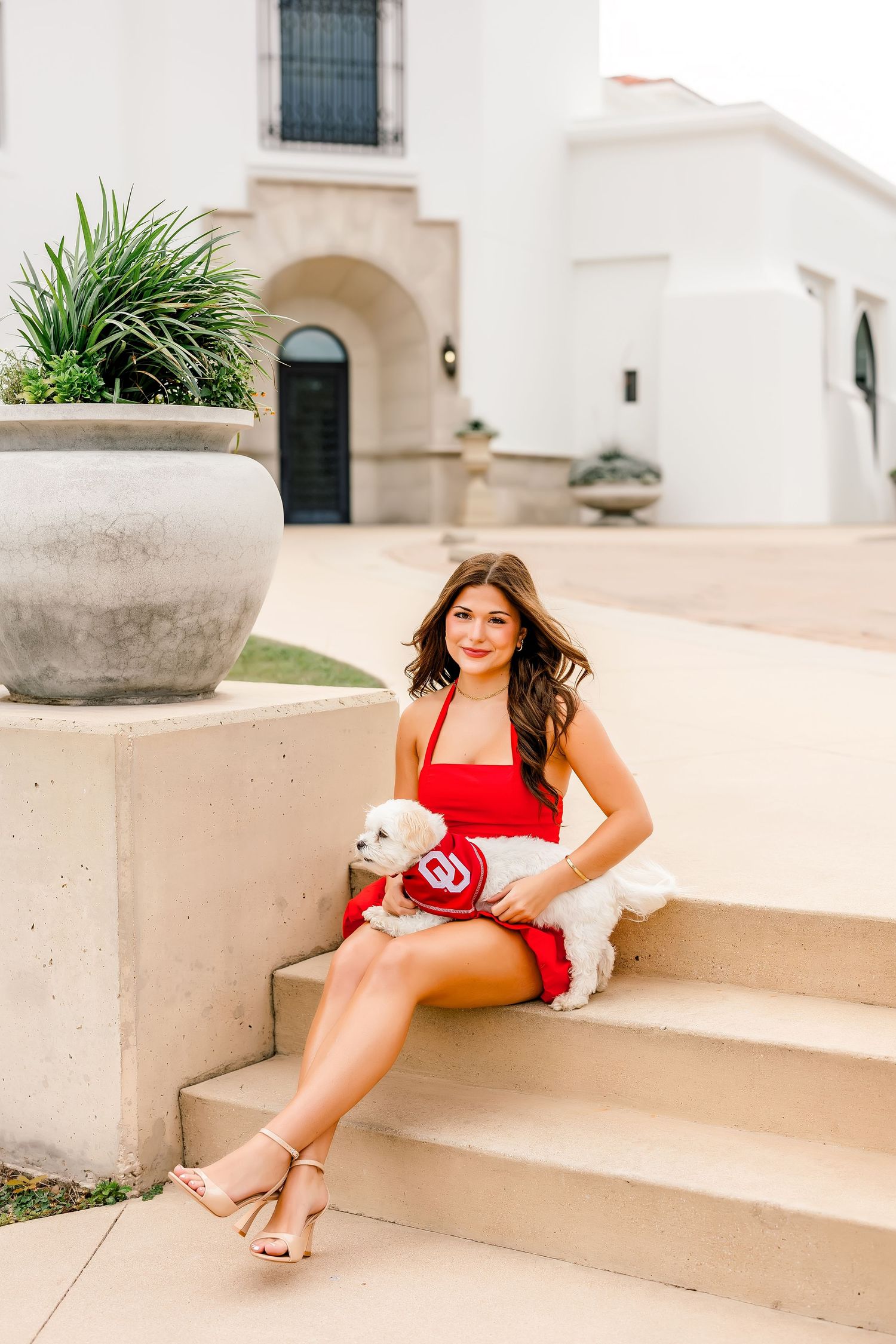 Young woman in red dress holding white dog with OU logo sitting on outdoor steps.