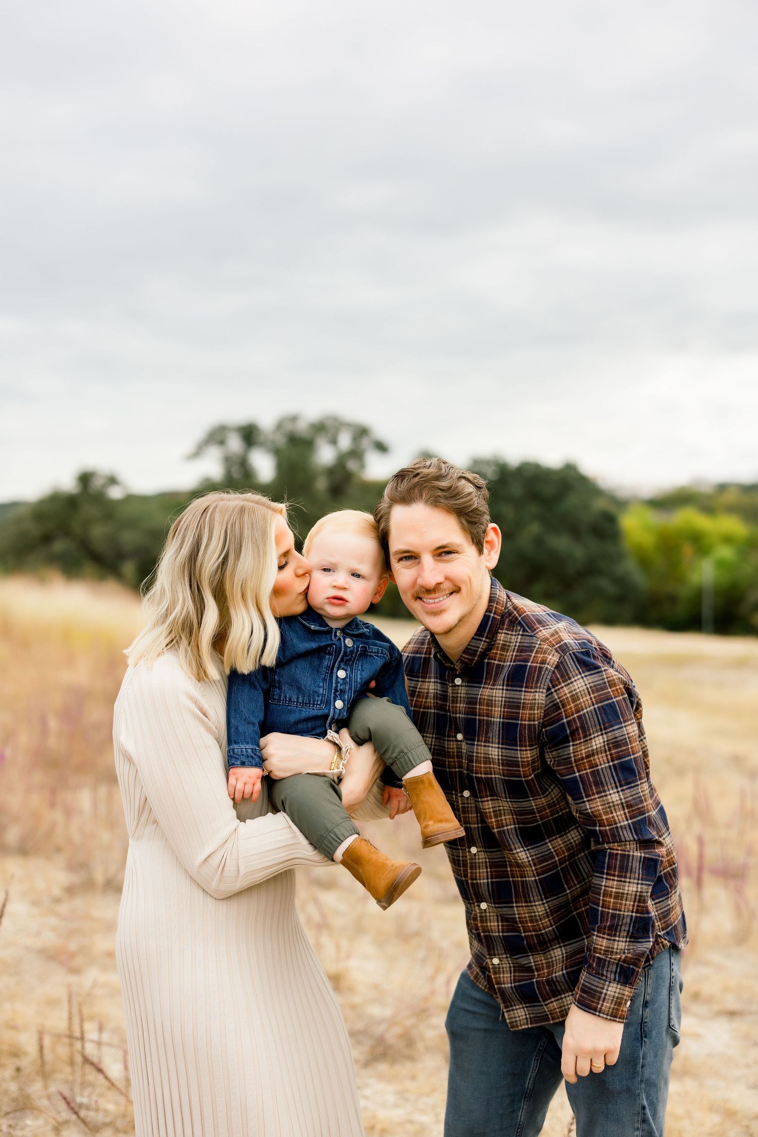 Mother kissing baby boy on cheek while father smiles in a golden dry grass field outdoor morning mini session at Bee Cave Park in Bee Cave.