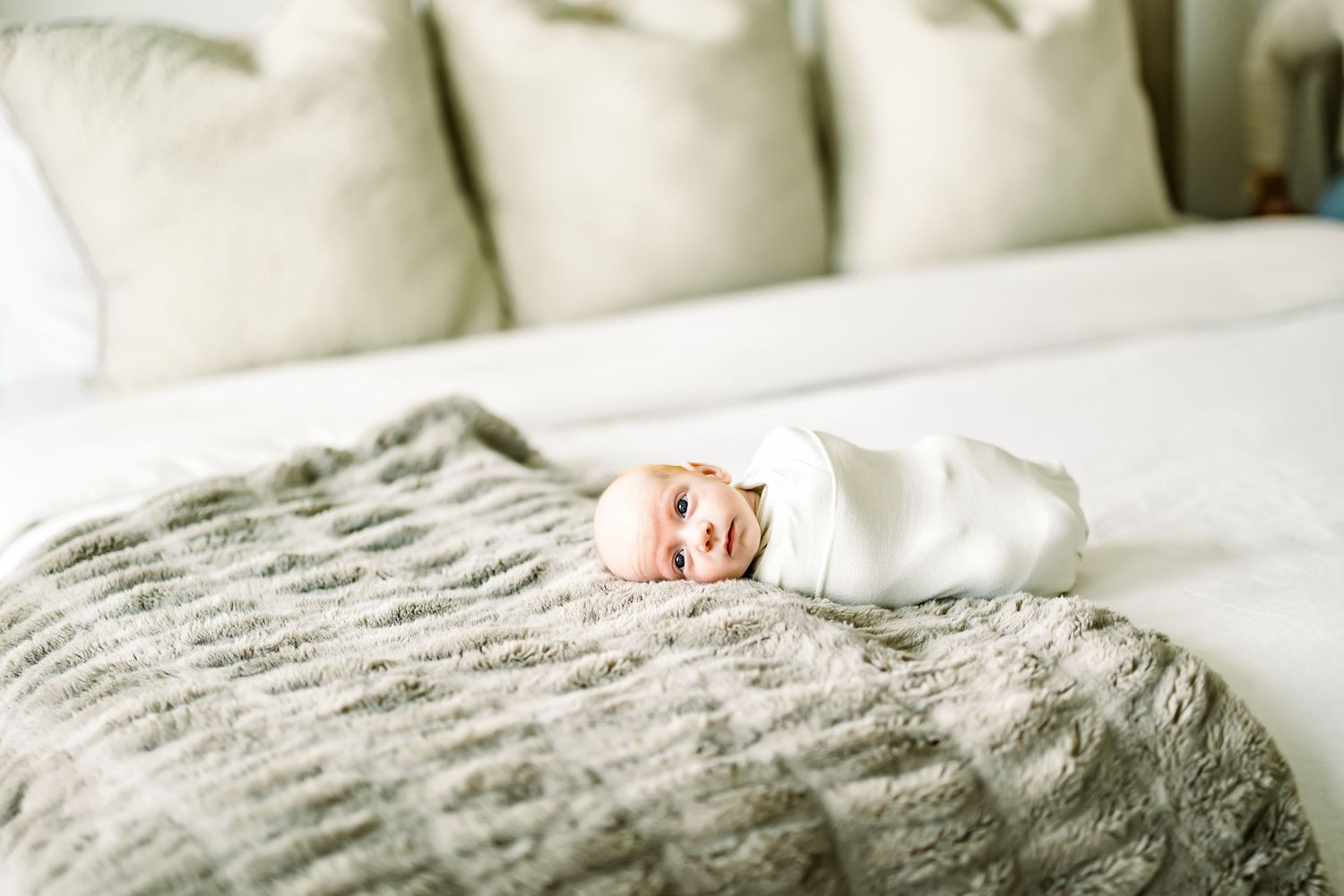 Newborn baby swaddled in white wrap smiling on a white bed with a gray textured blanket and cream pillows.