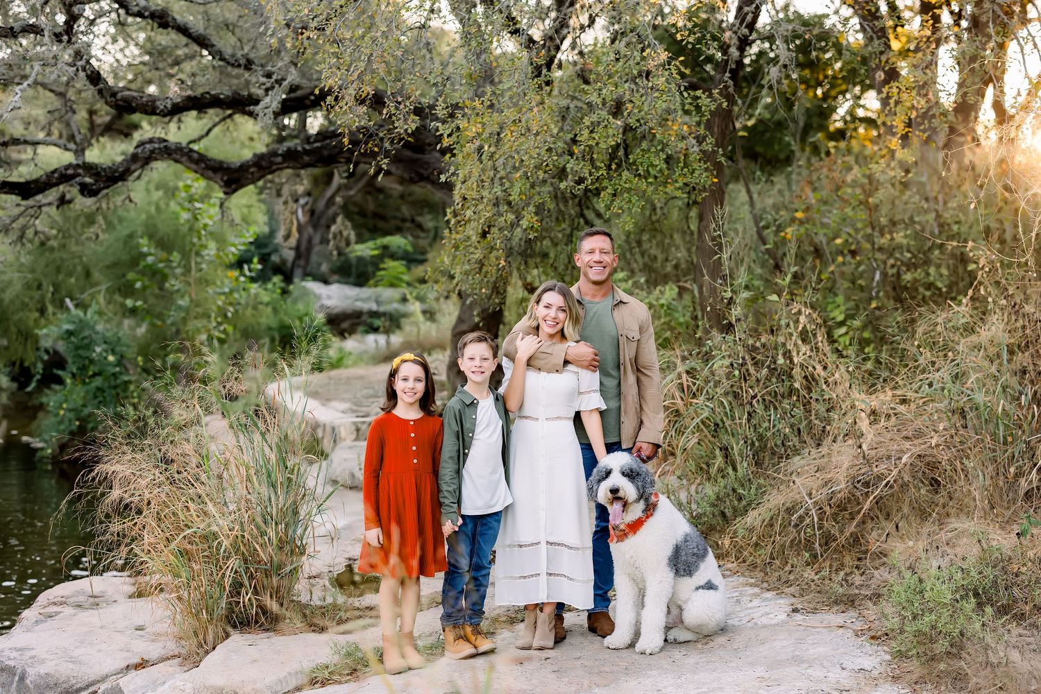 Family with three kids and large dog posing on rocks beside creek in colorful fall outfits.