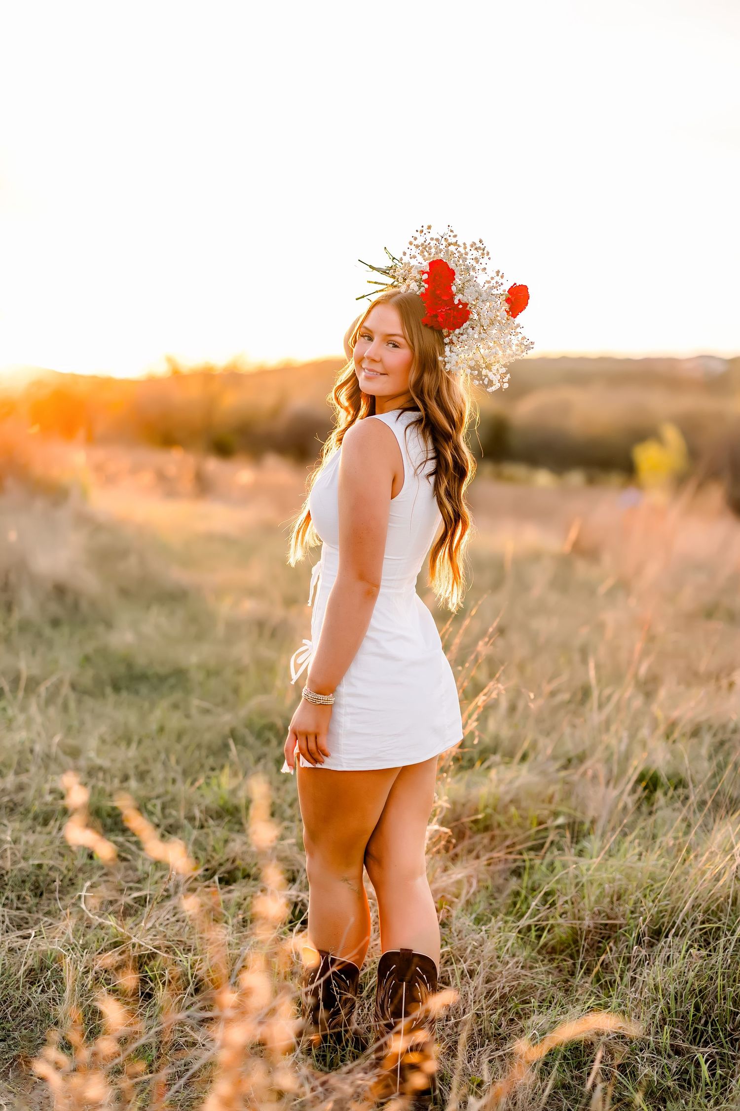 A young woman in a white dress with a floral crown poses in a golden sunset field.