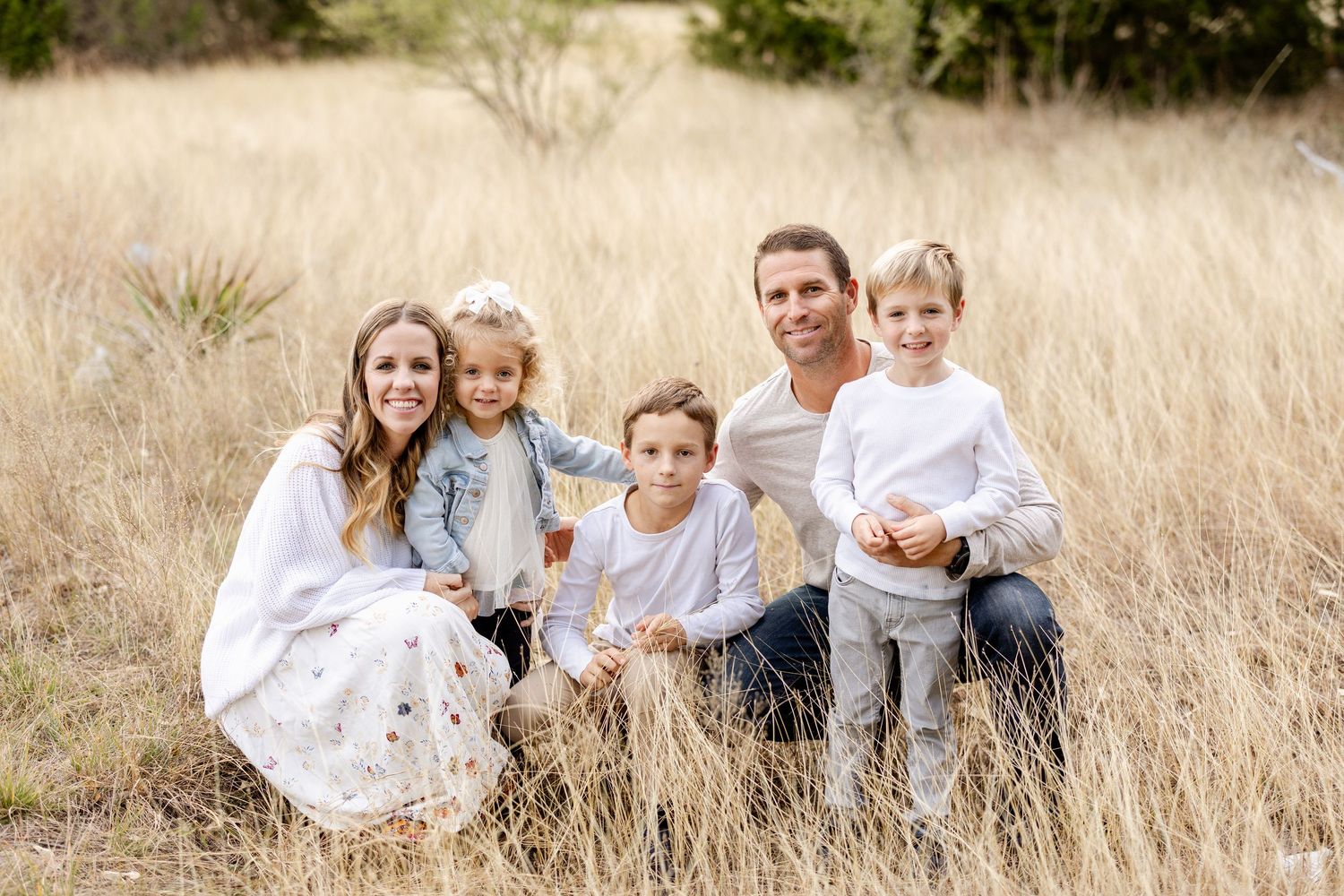 A smiling family of five poses together in tall golden grass during a bright outdoor portrait session in nature.