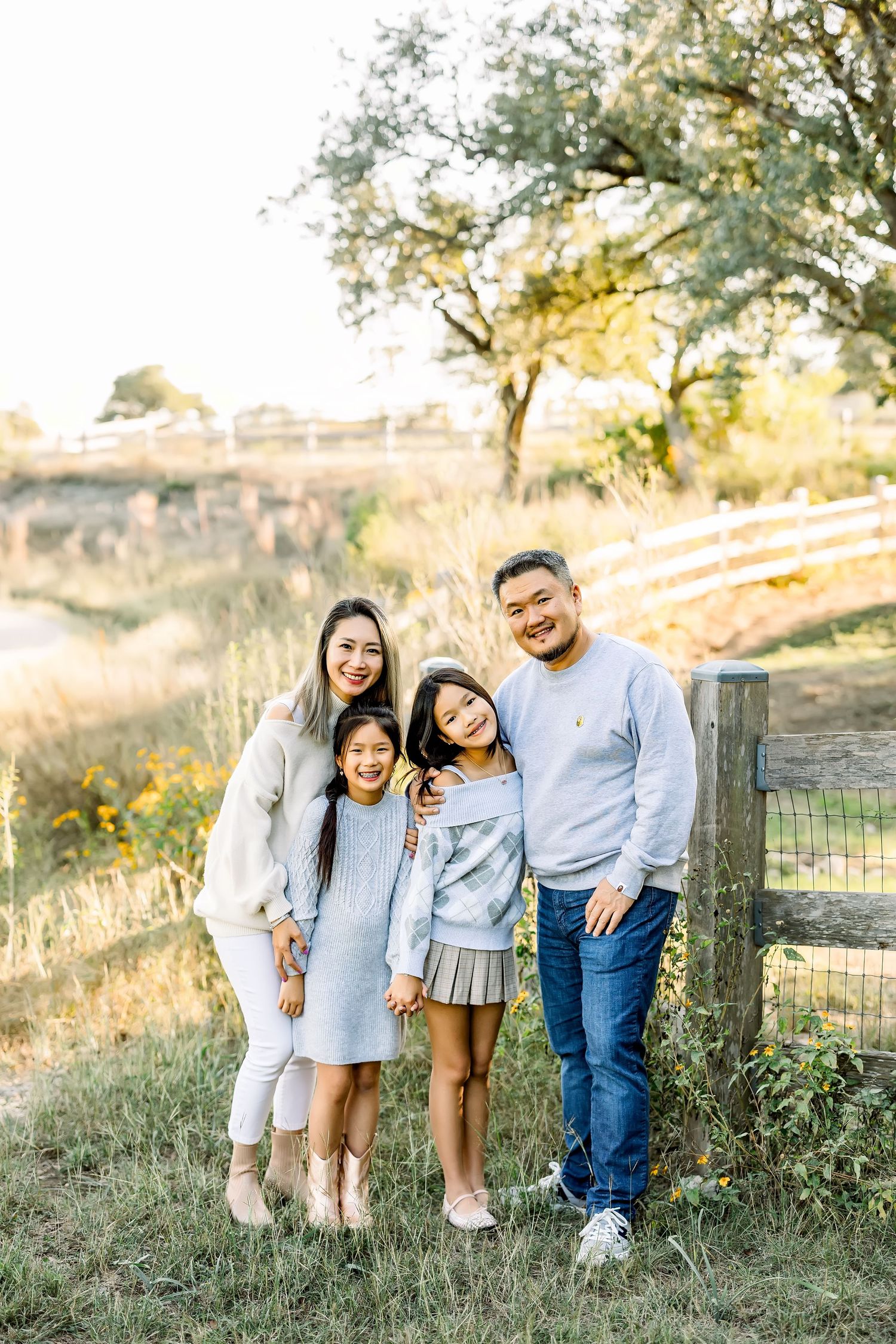 Asian family of four smiling together near a wooden fence outdoors at a mini session in Bee Cave, Tx  by Shannon Worley Photography.