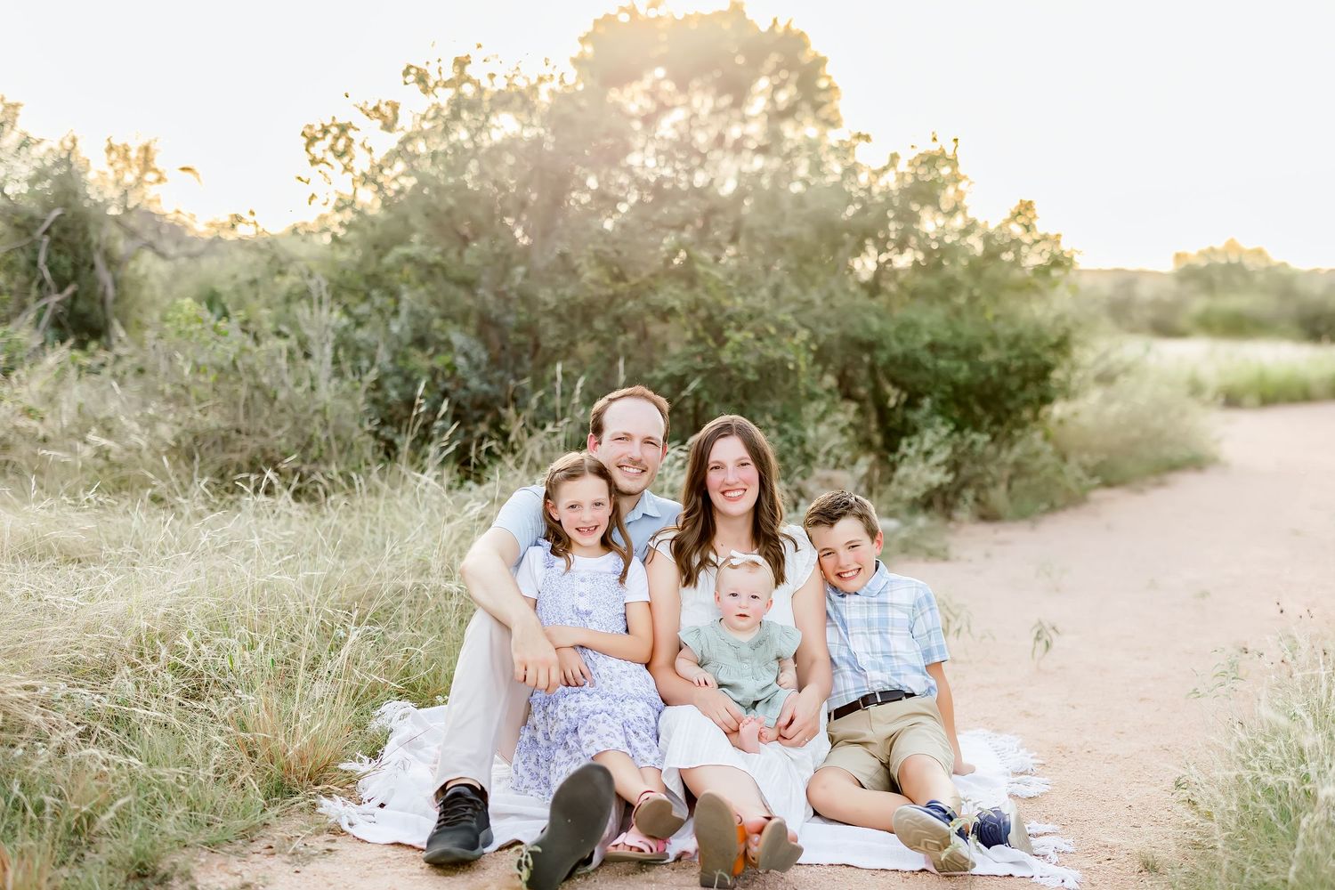 Family of five sitting together on sandy path surrounded by golden brush, smiling warmly at camera.