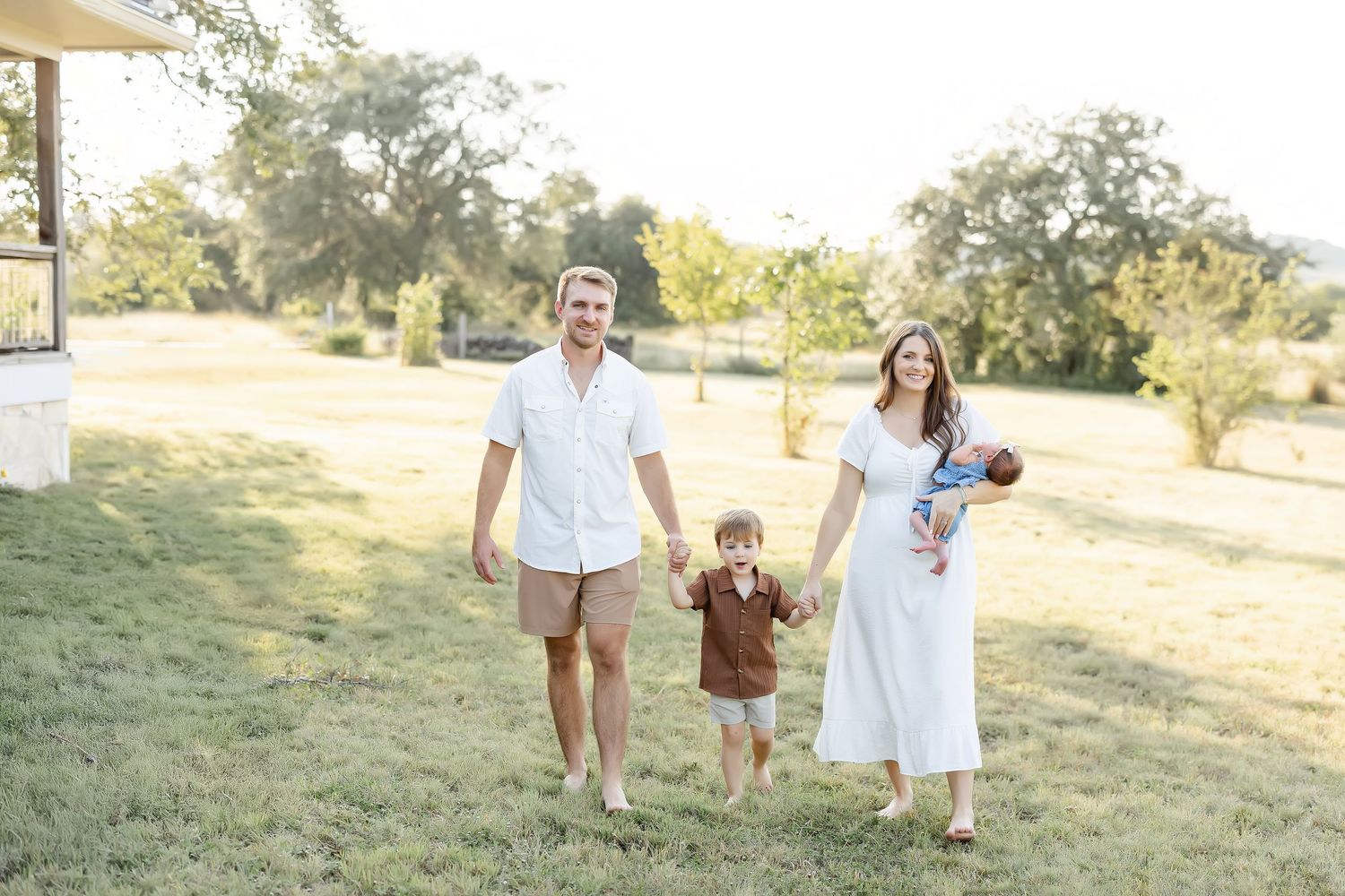 A happy family of four walks outdoors on a sunny day, parents holding hands with toddler while mom carries newborn baby.