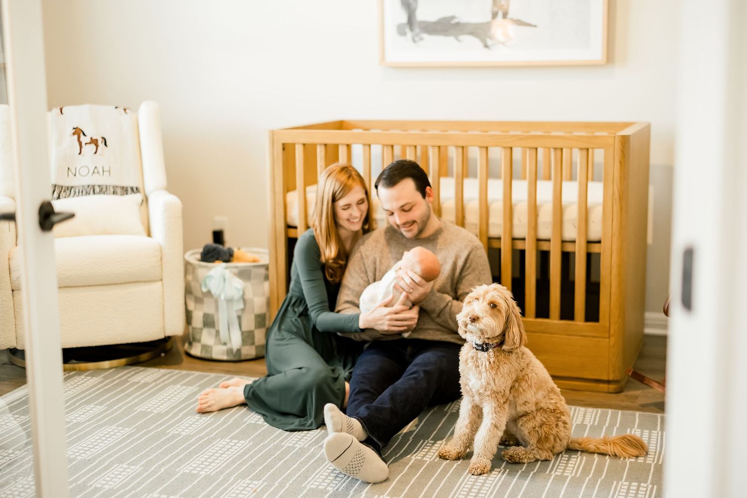 A couple sits on a nursery floor with their newborn baby and golden doodle dog in front of a wooden crib.