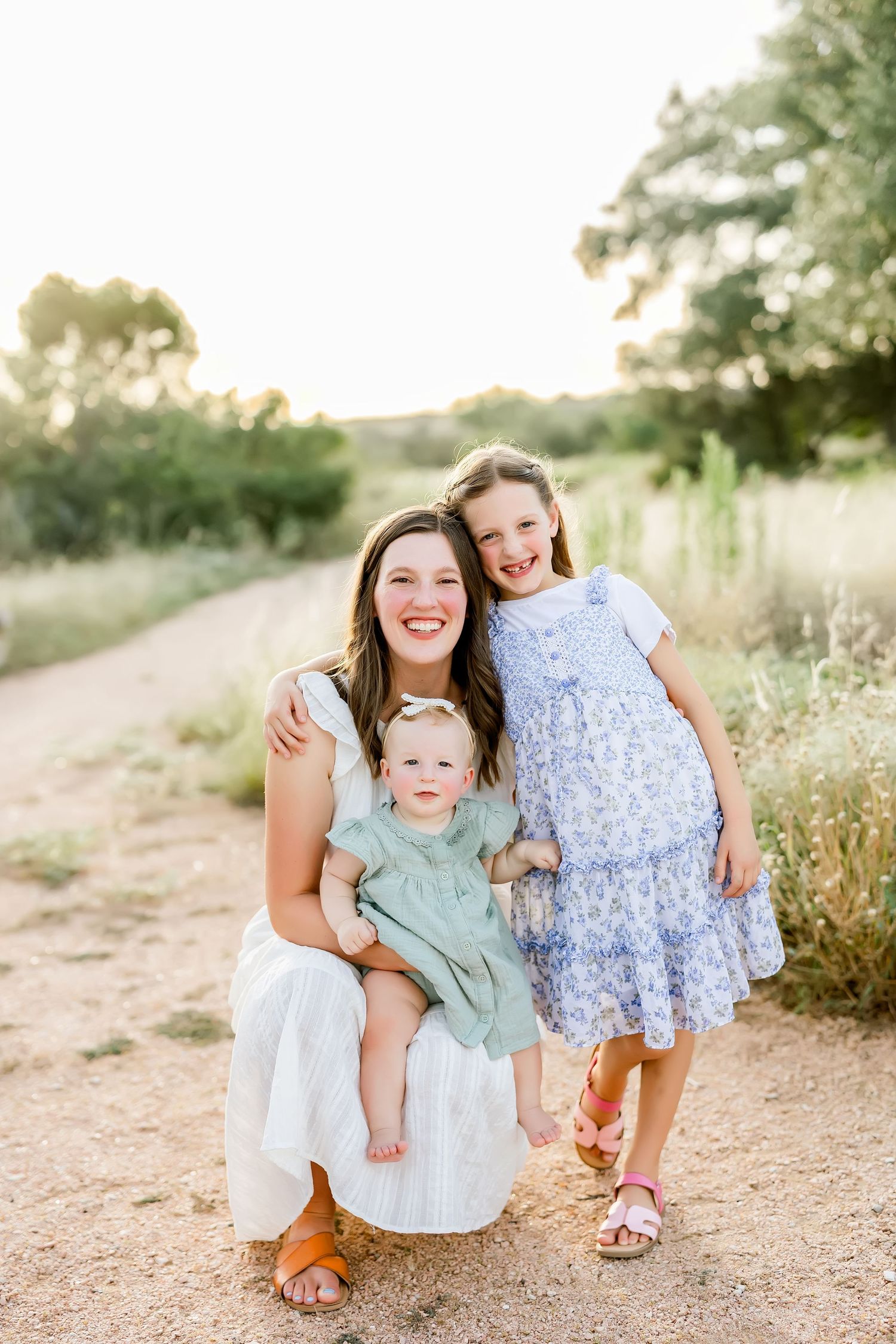 Mother crouches on dirt path holding baby while older daughter stands beside her at sunset.