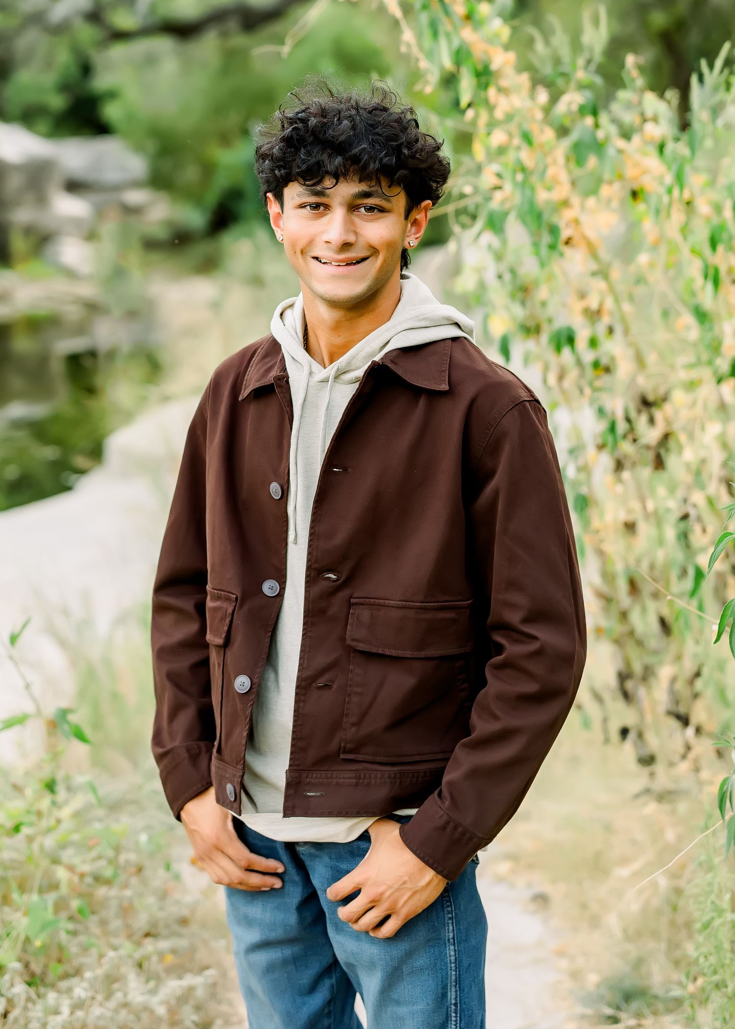 Smiling young man with curly hair wearing a brown jacket and jeans poses outdoors near green plants.