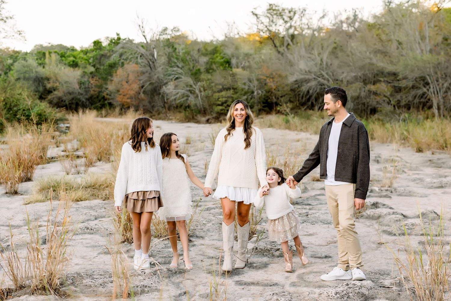 Family walking hand in hand through sandy field in coordinating white and neutral outfits.